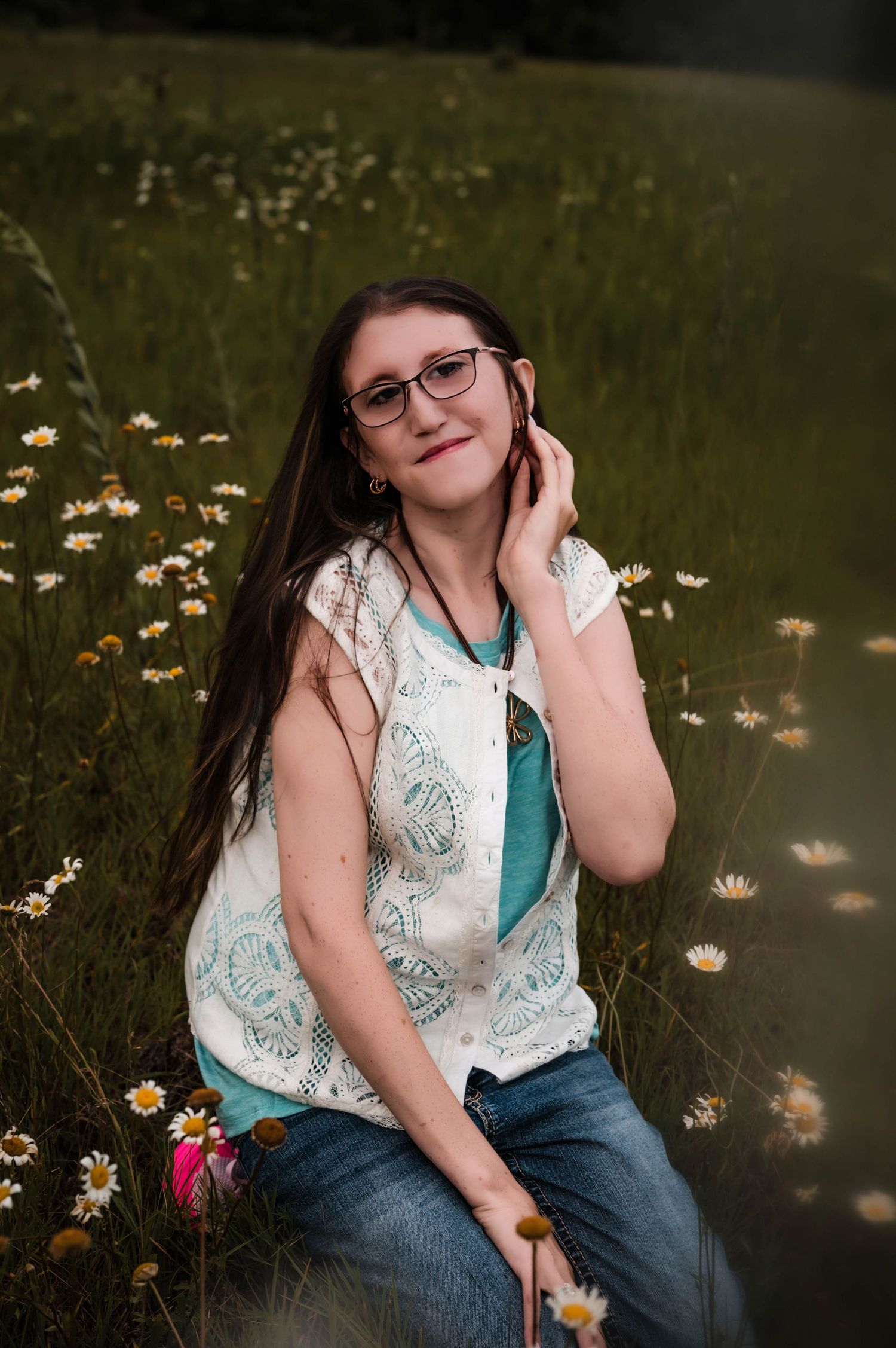 Young woman with glasses sitting in a field of daisies, wearing jeans and a white lace vest over a teal top.