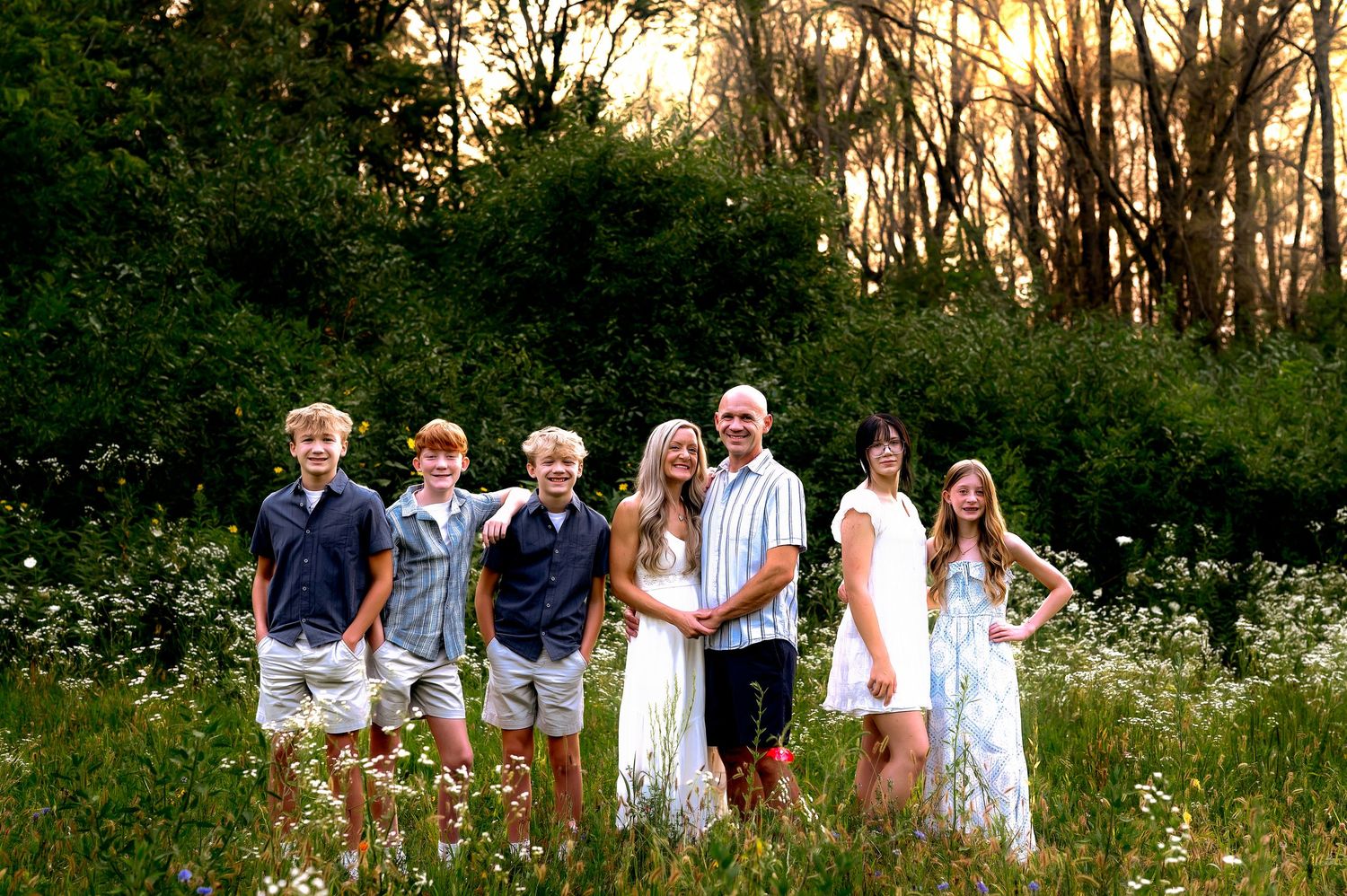 A family group poses together in a wildflower meadow at sunset with trees in the background at a private wildflower field, in Kewaskum, WI