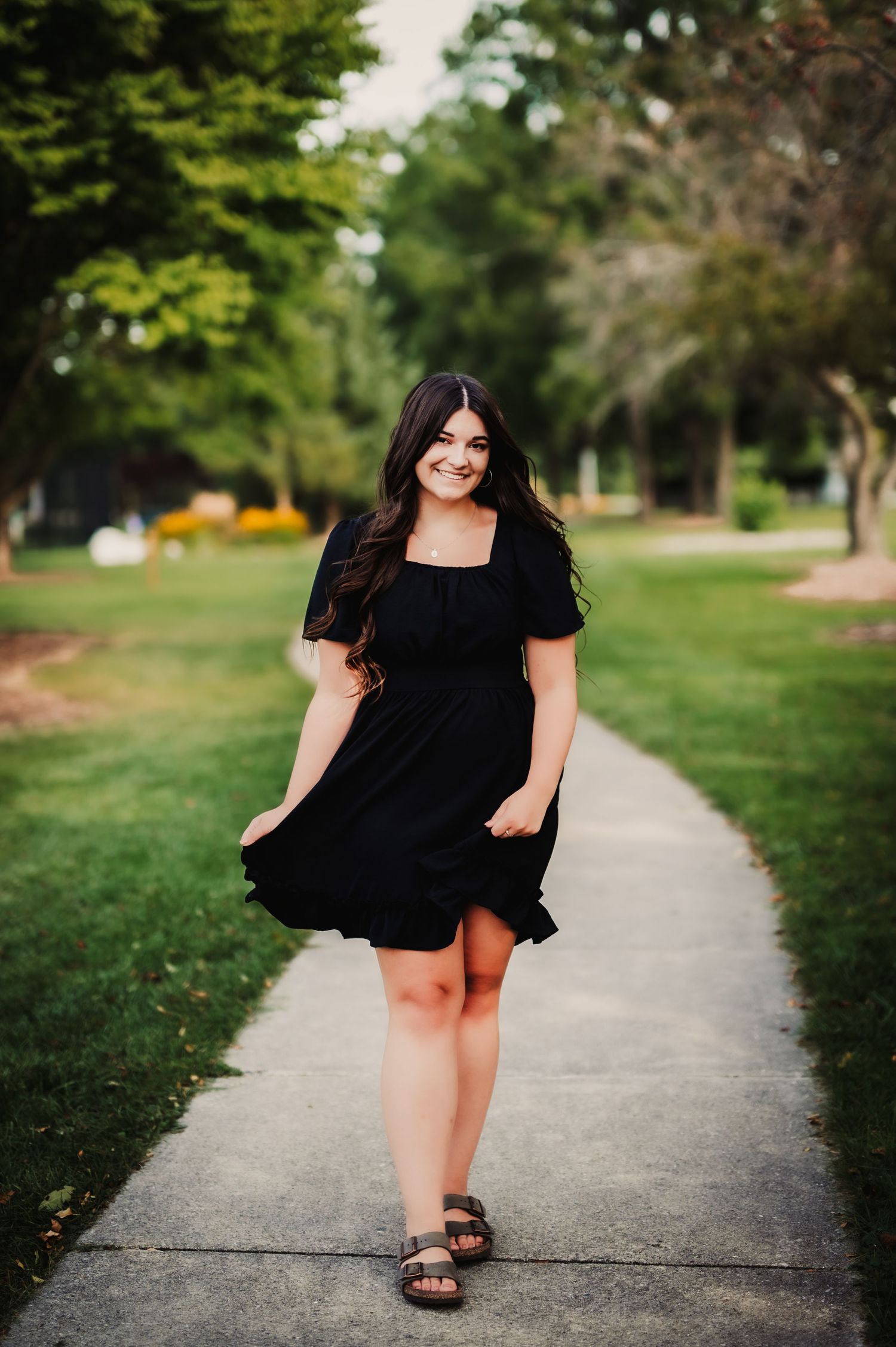 Figure in flowing black dress walks along a concrete path surrounded by green trees.
