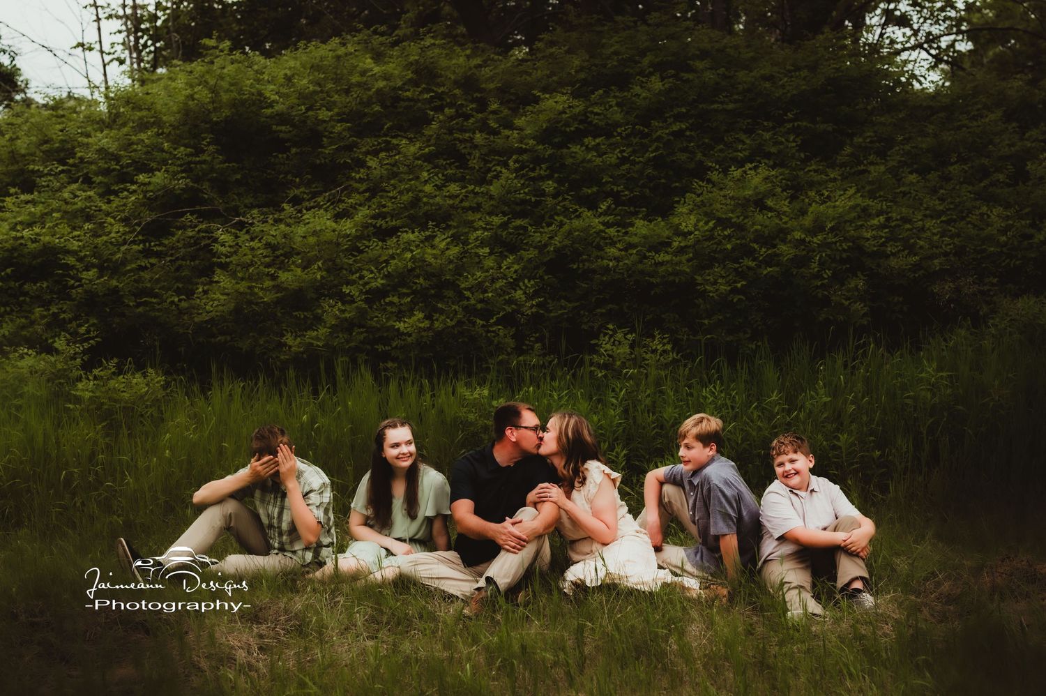 Family group sits casually on grassy hill for relaxed outdoor portrait session.