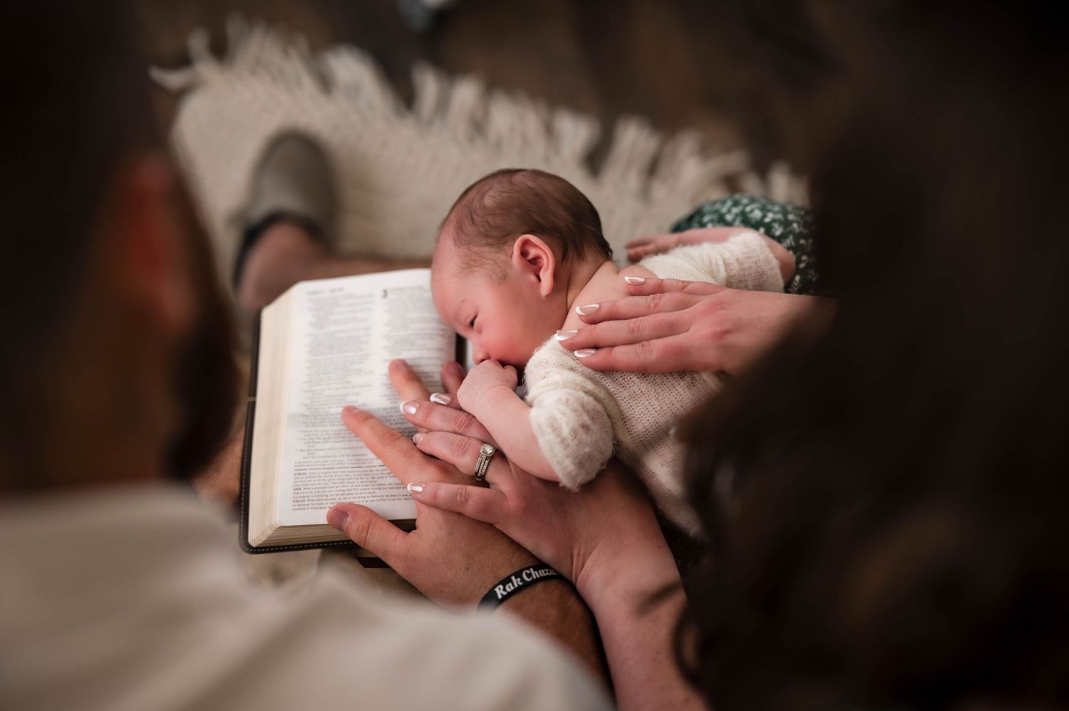 Parents' hands hold an open Bible while a sleeping newborn baby rests against it in a warm, intimate family moment.