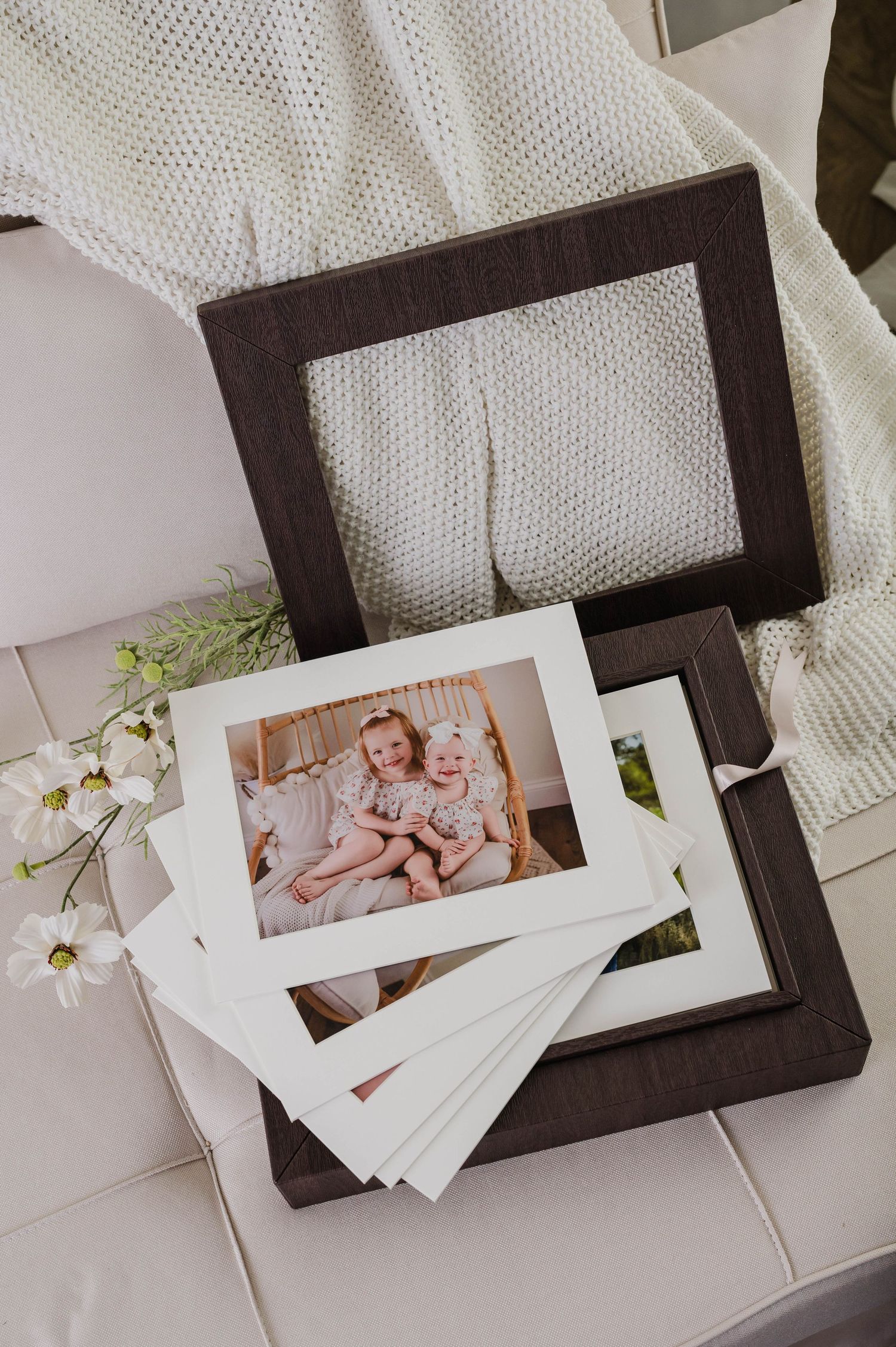 Matted family photos of two young children stacked on dark wood frames atop a white tufted sofa with a knit blanket.