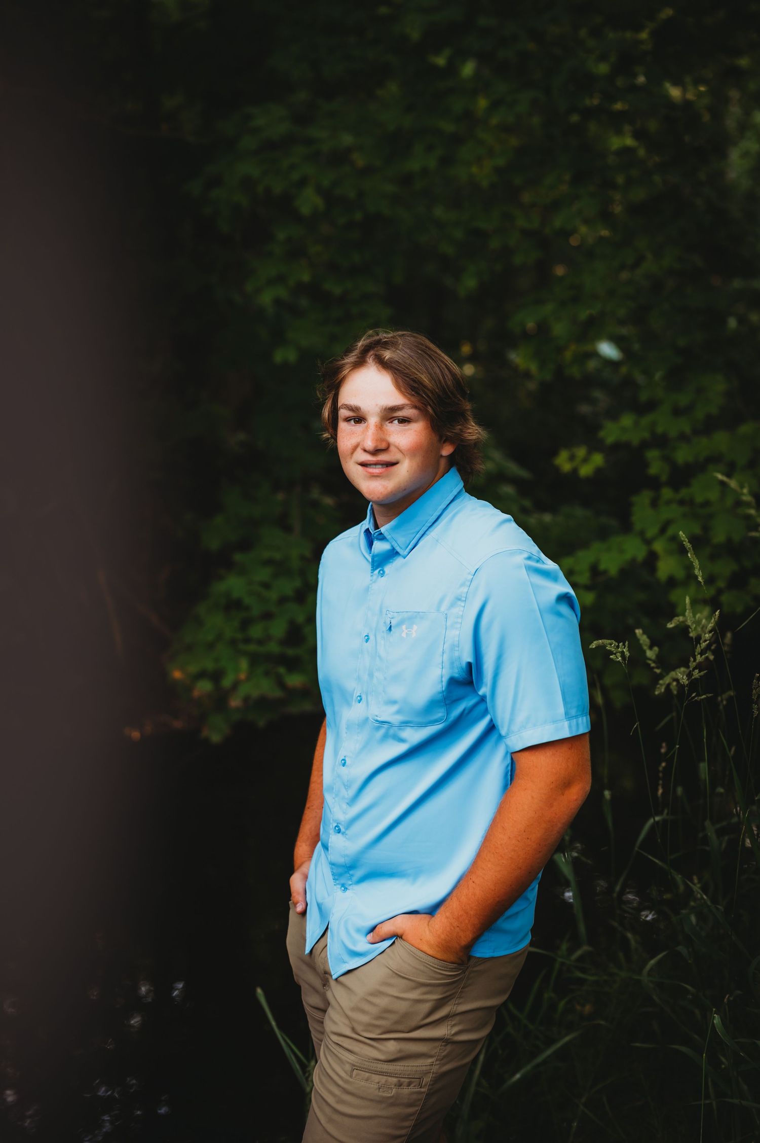 Young person in light blue polo and khaki shorts poses casually in wooded outdoor setting.