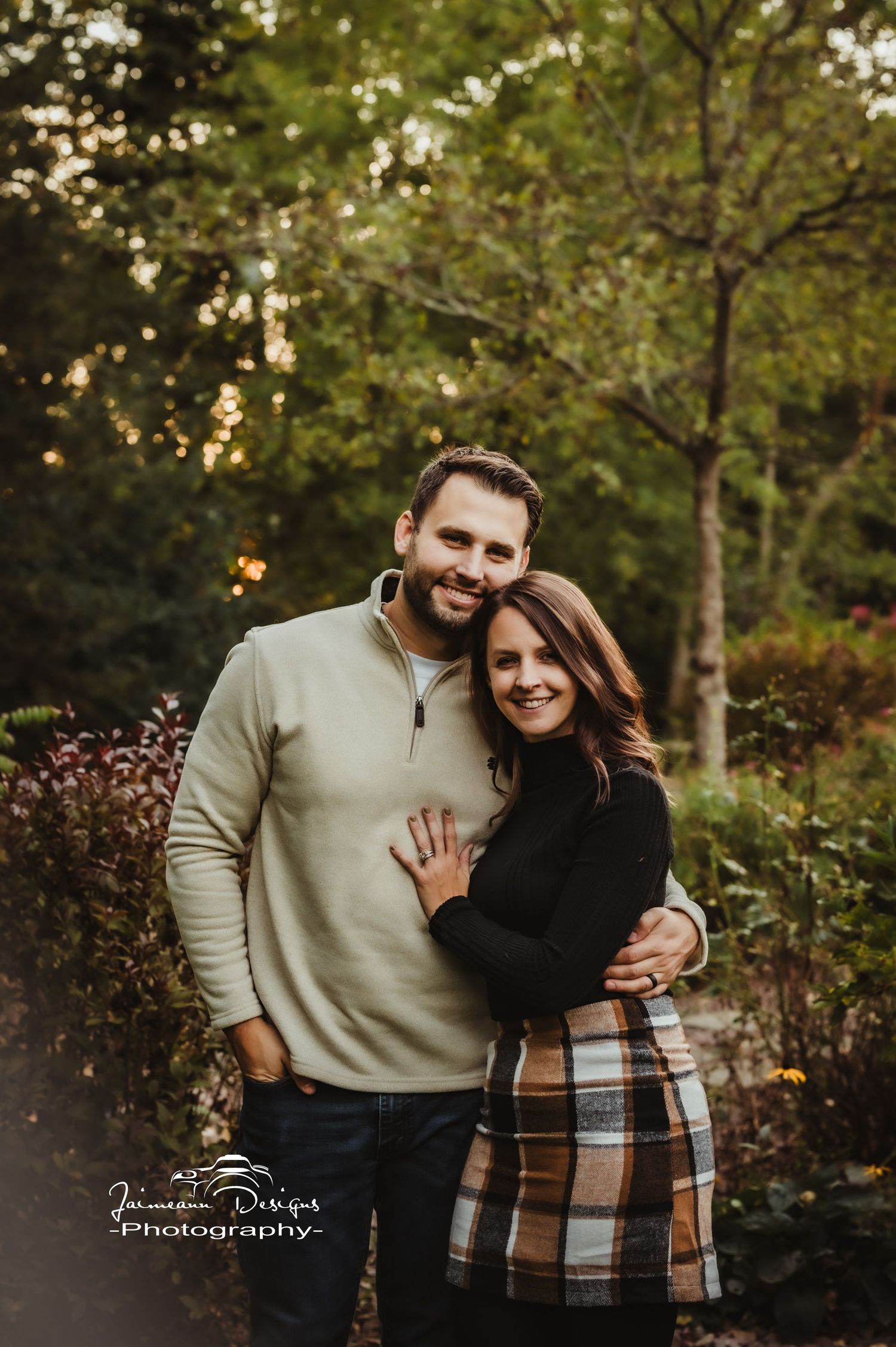 A couple embraces outdoors wearing casual sweaters against a backdrop of fall foliage.