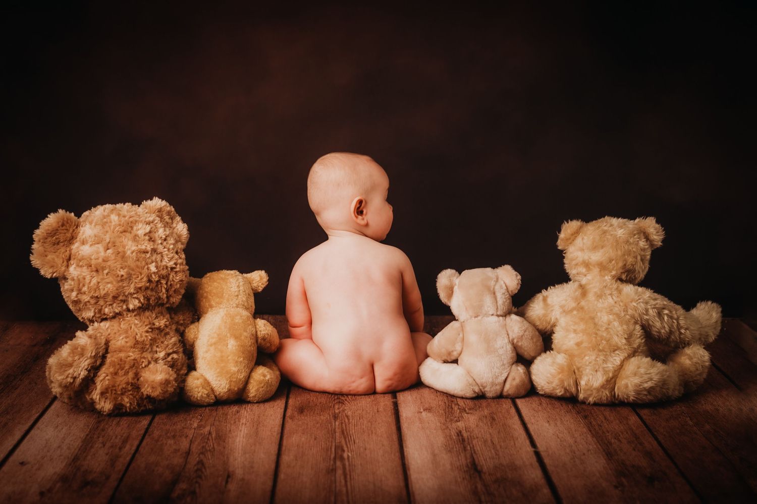 A naked baby sits among teddy bears on wooden flooring against a dark background.