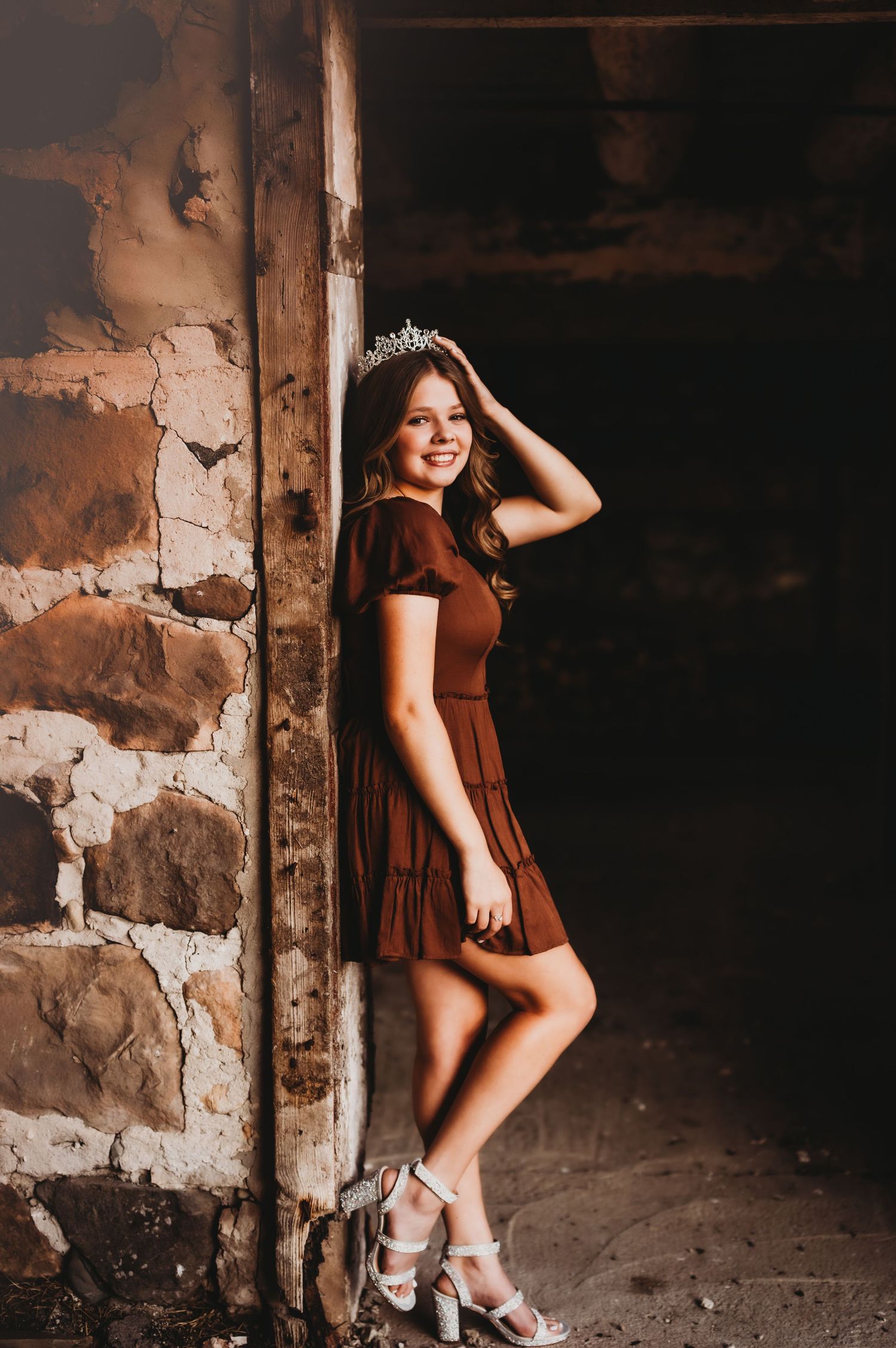 Woman in brown dress poses against rustic stone wall in moody lighting. Senior Prom Queen, WI