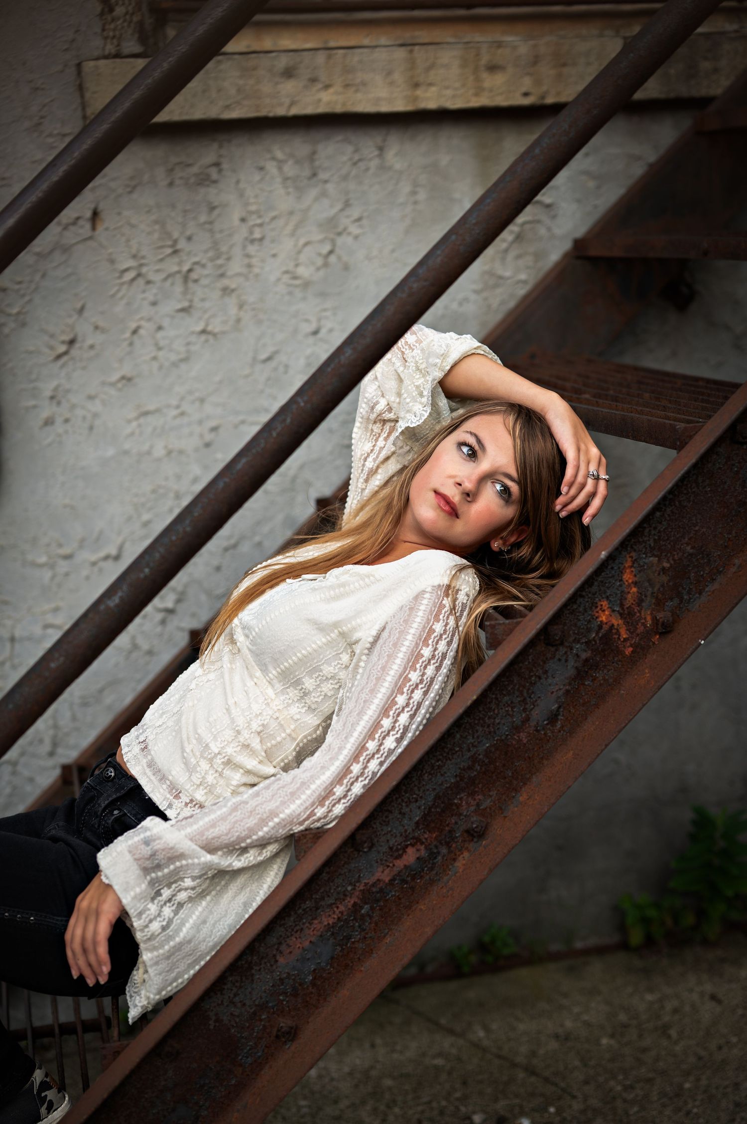 A woman in a white lace top poses artistically on rustic metal stairs in an outdoor setting in Kewaskum, WI