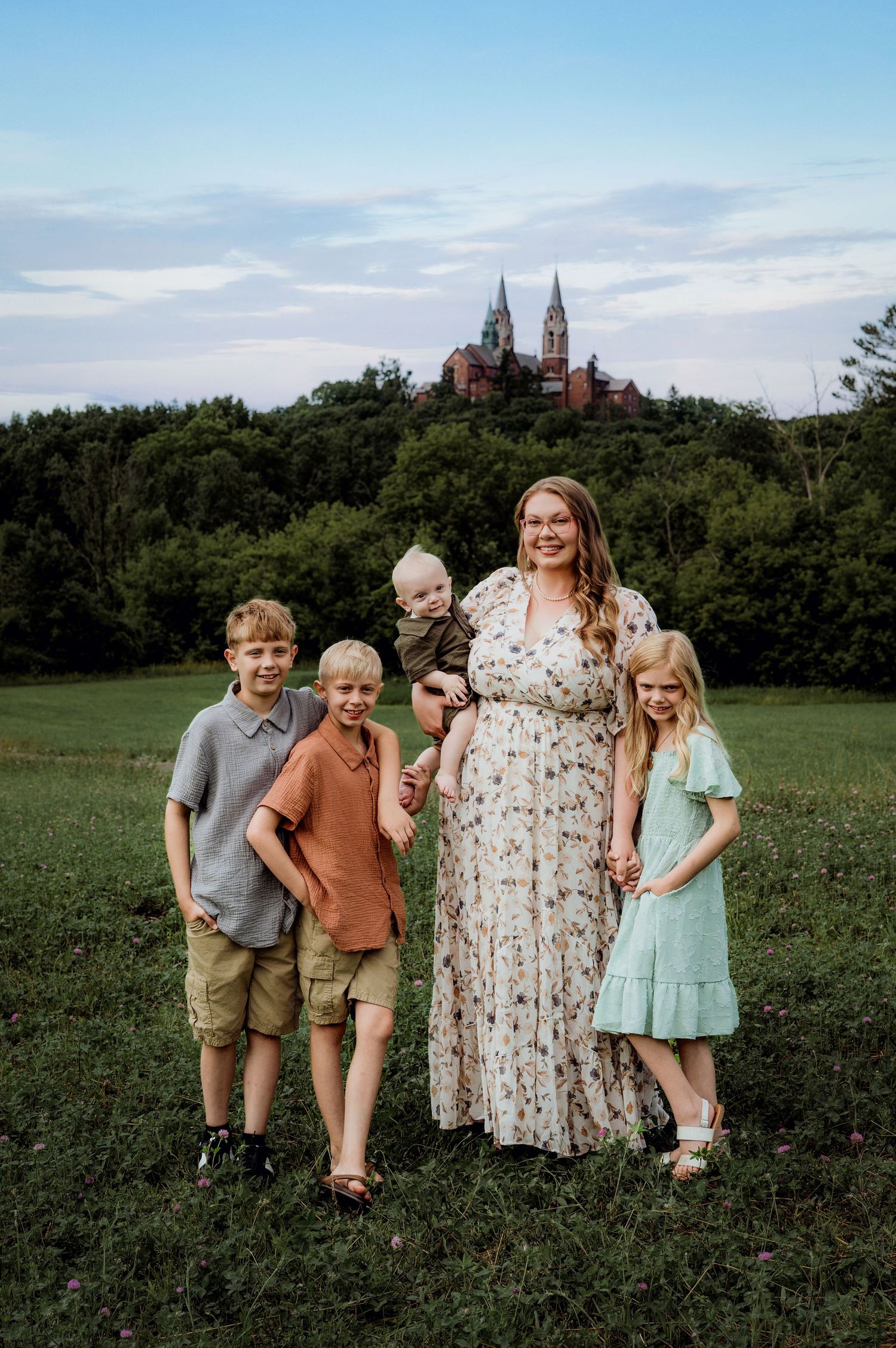 A mother in a floral dress poses with her four children in a green field with a historic castle on a hill behind them.