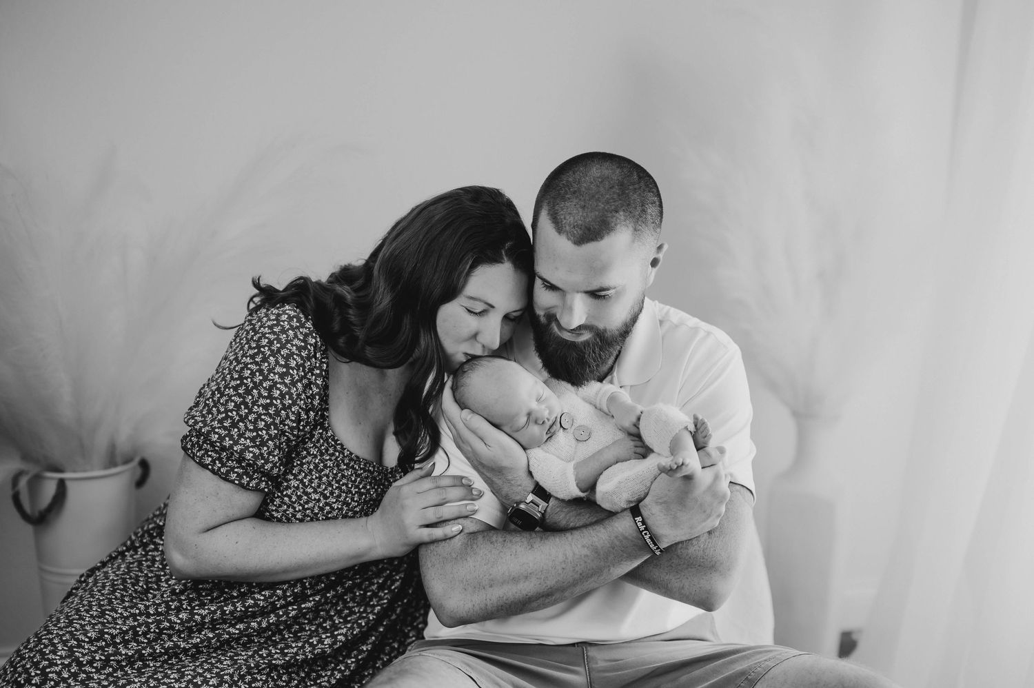 Black and white newborn family portrait of a mother and bearded father tenderly cradling and kissing their sleeping baby.
