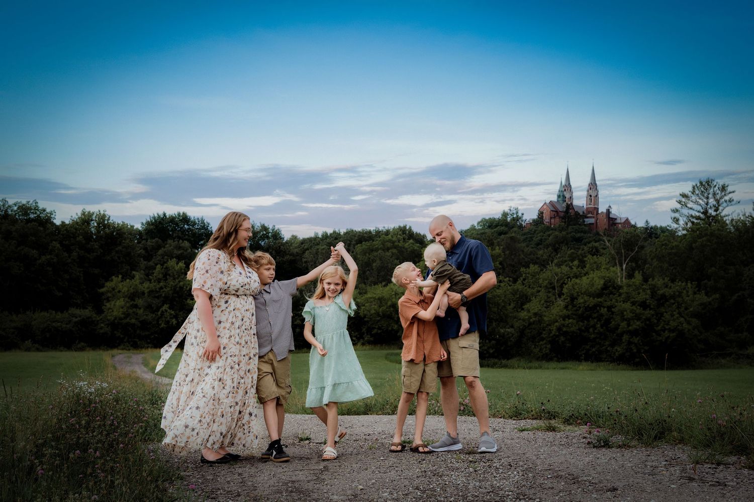 Family of five dancing outdoors on a gravel path with lush trees and a church spire at dusk.