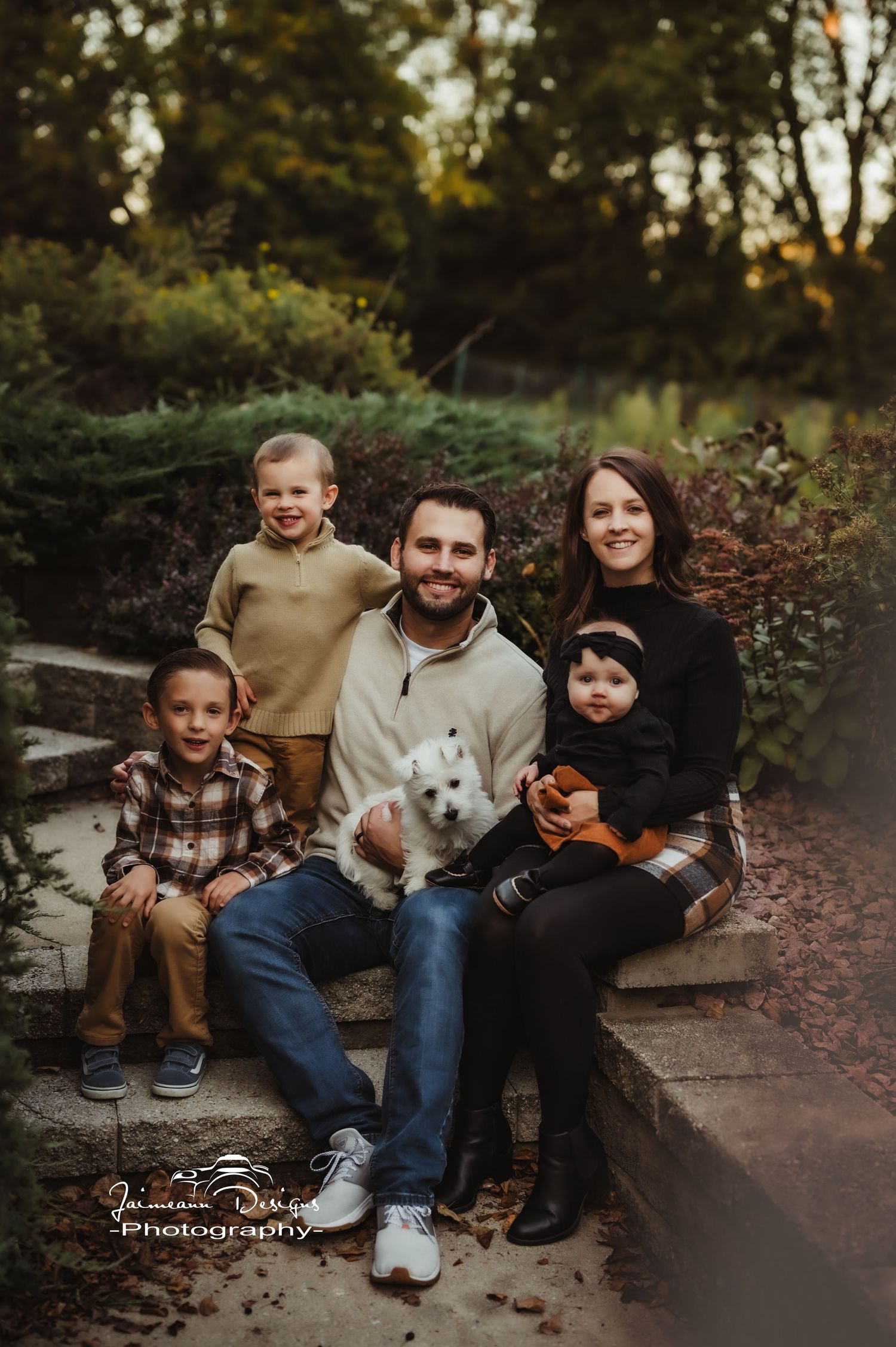 A family with three children and a small white dog sit together on stone steps in an autumn garden setting.