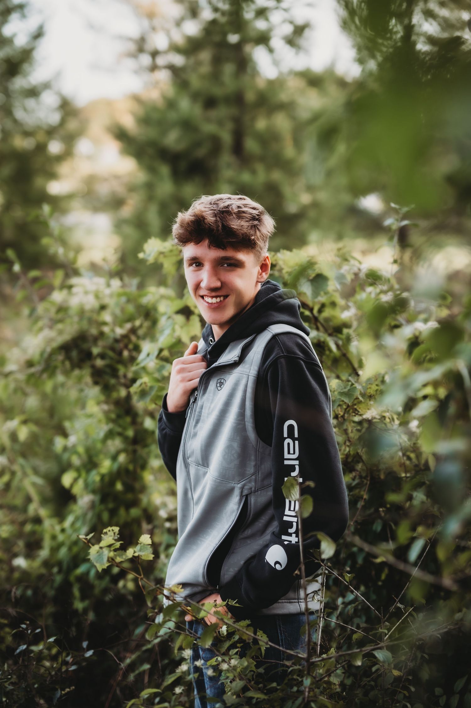 Person wearing grey and black hoodie standing among green foliage.