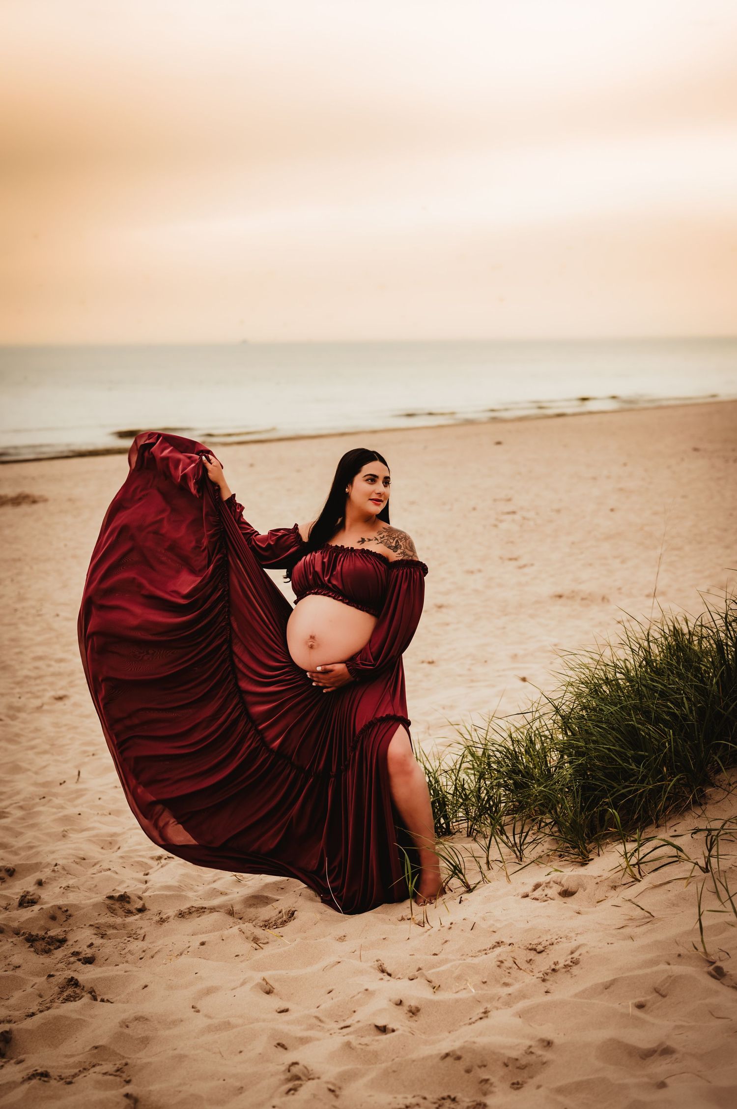 A flowing burgundy dress billows dramatically against sandy dunes at sunset on a peaceful beach.