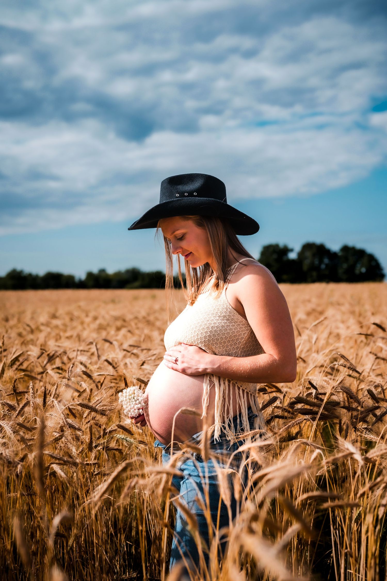Pregnant woman in black cowboy hat cradling baby bump in golden wheat field under blue sky.