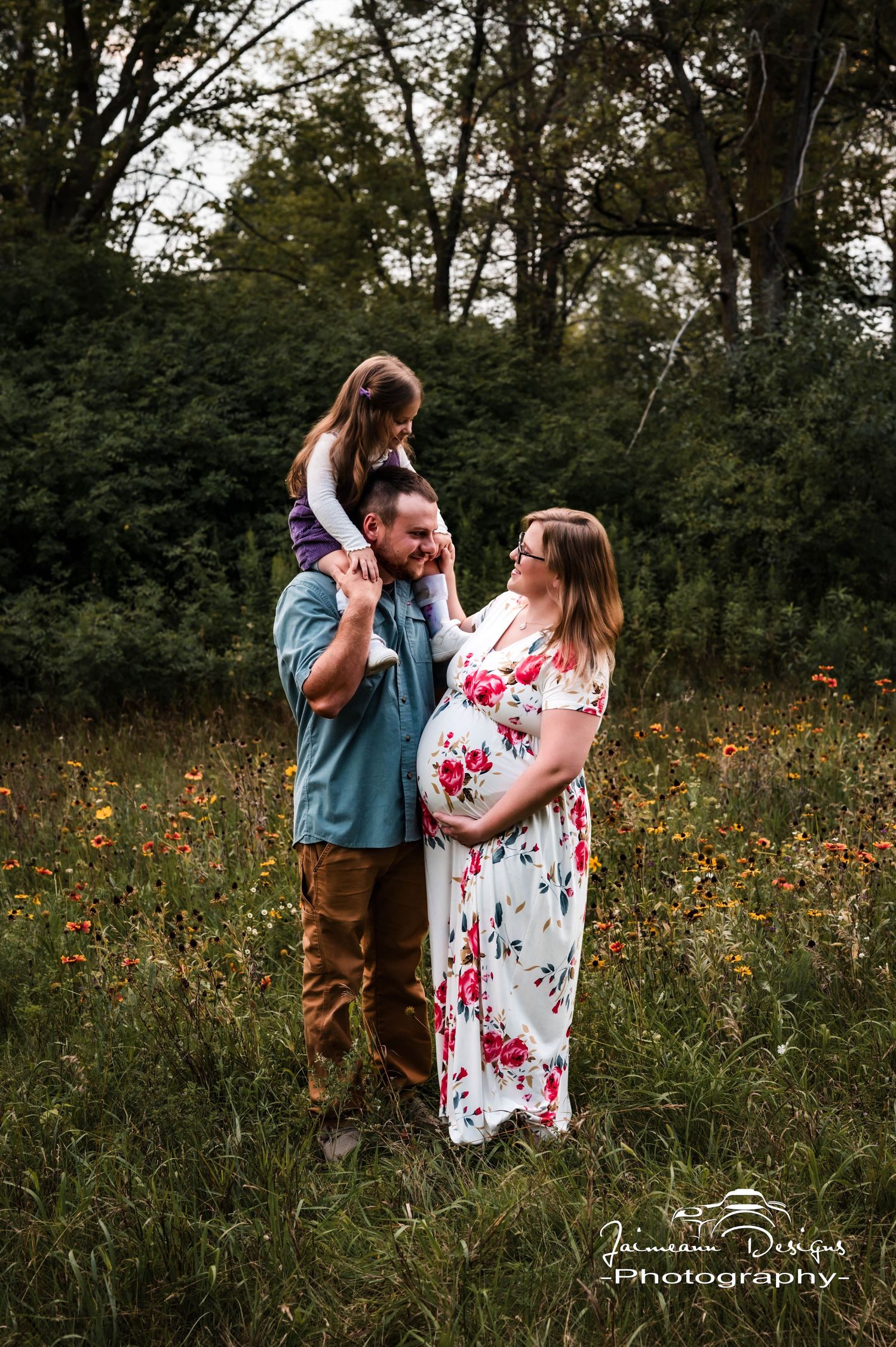 Pregnant woman in floral dress with husband and daughter on his shoulders posing in a wildflower field for maternity photos.