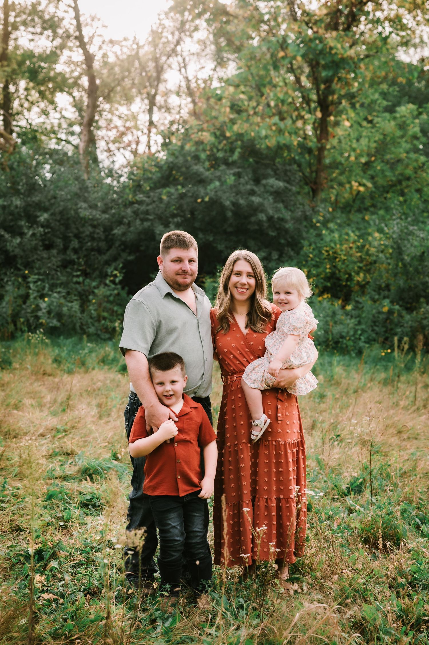 Family of four posing outdoors in a lush green field, parents with young boy and baby girl in warm autumn tones.