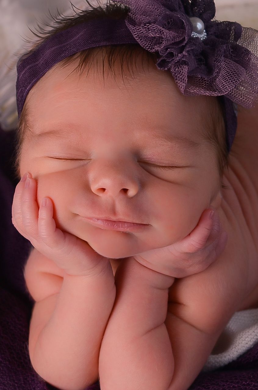 A sleeping baby wearing a purple bow headband rests peacefully in soft lighting.