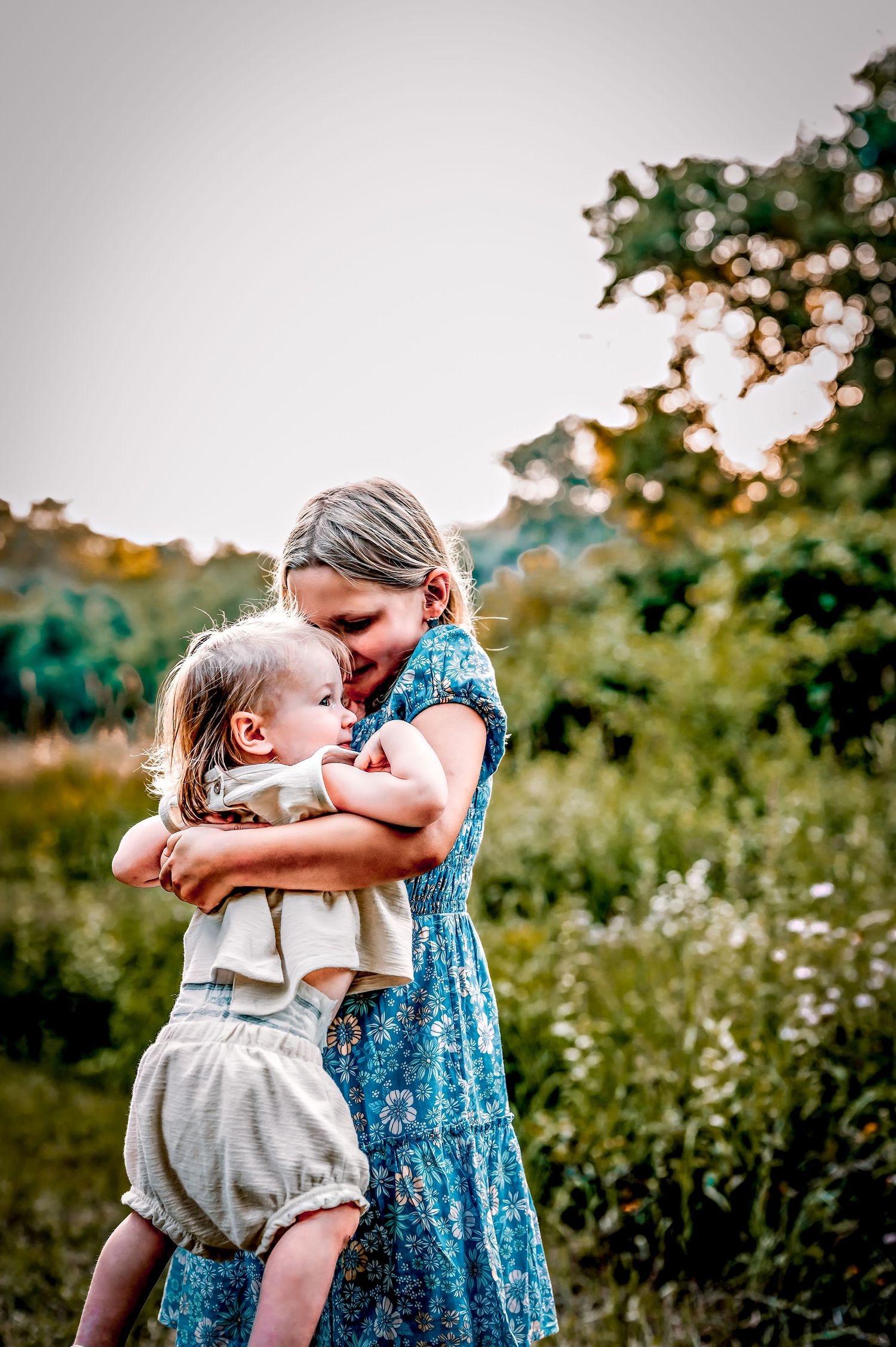 Two young children in casual summer outfits share a sweet moment together in a sunlit garden with wildflowers.