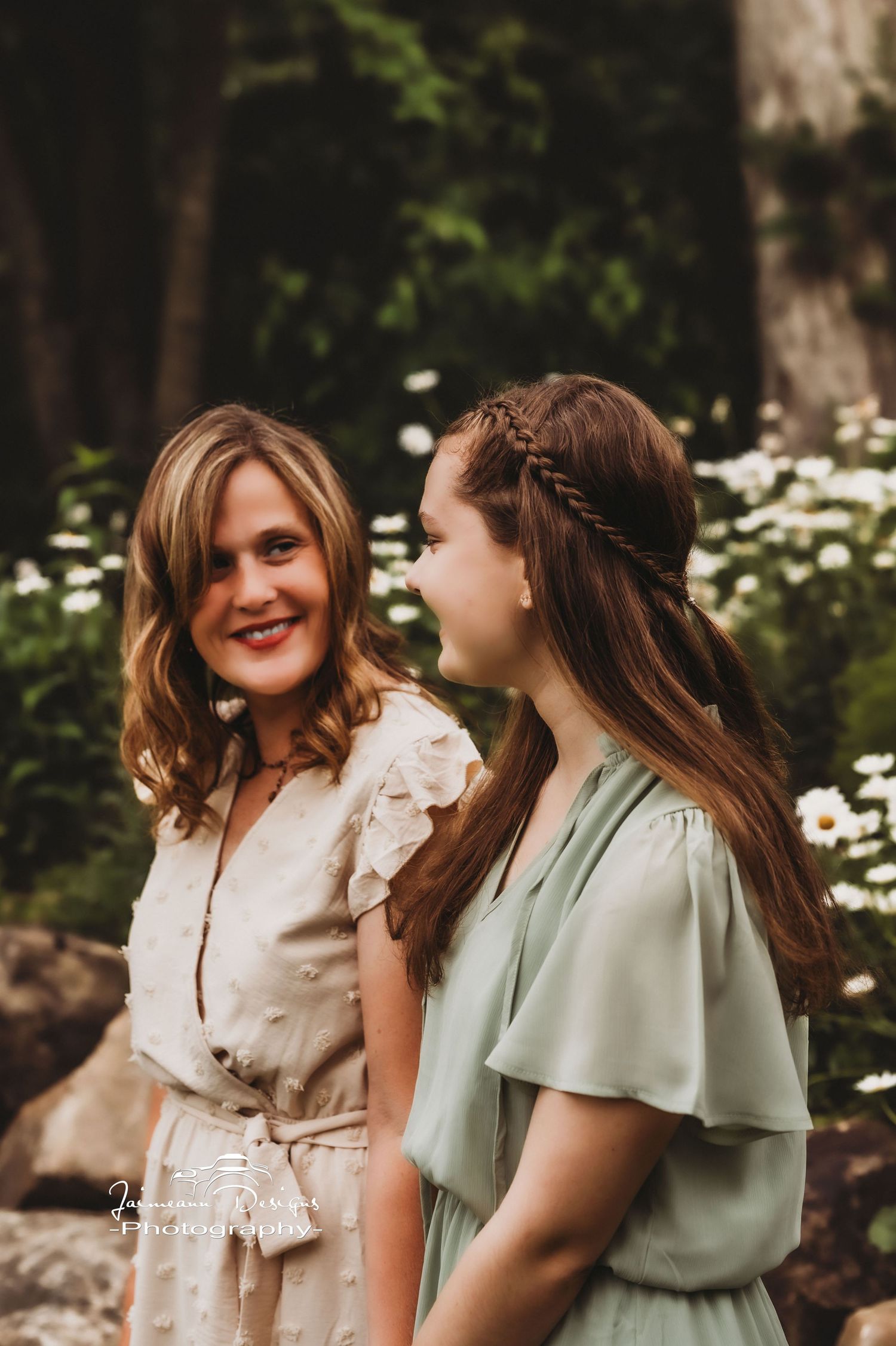 Two people in casual dresses share a candid moment among blooming daisies in a wooded outdoor setting.