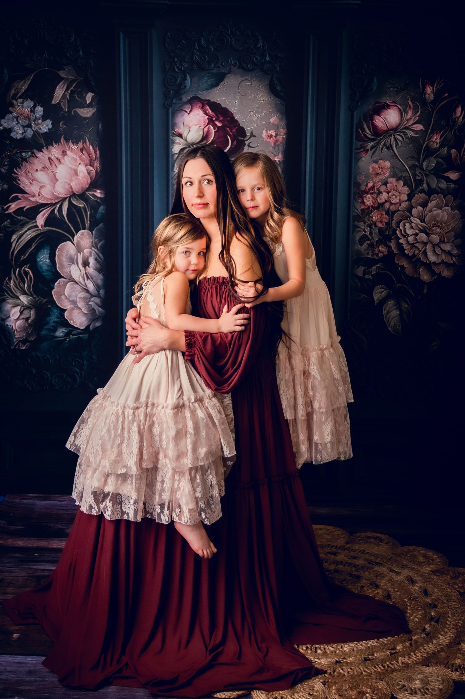 Elegant portrait with burgundy gown and cream lace dresses against dark floral backdrop.