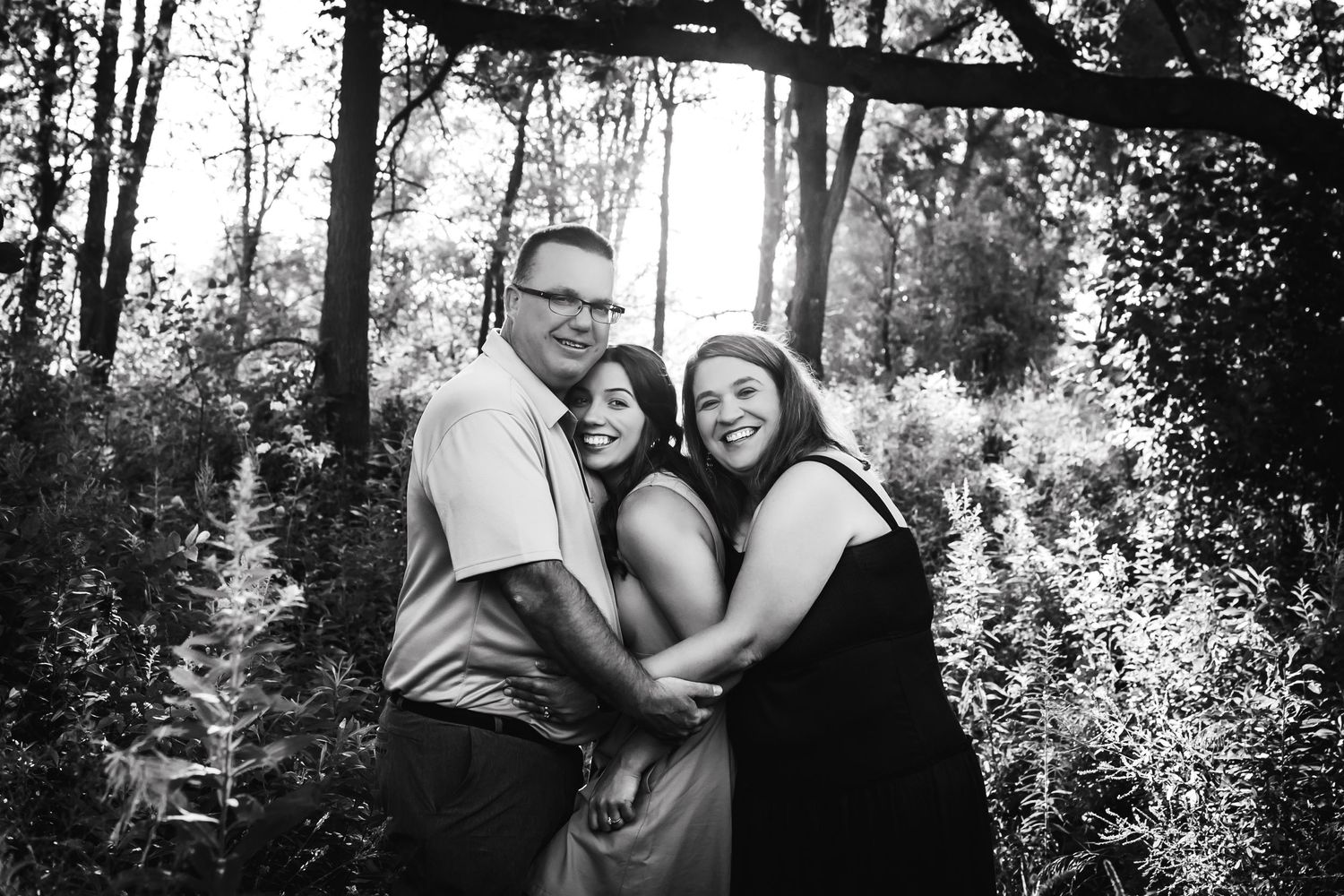 Black and white photo of three people embracing and laughing together in a wooded setting.
