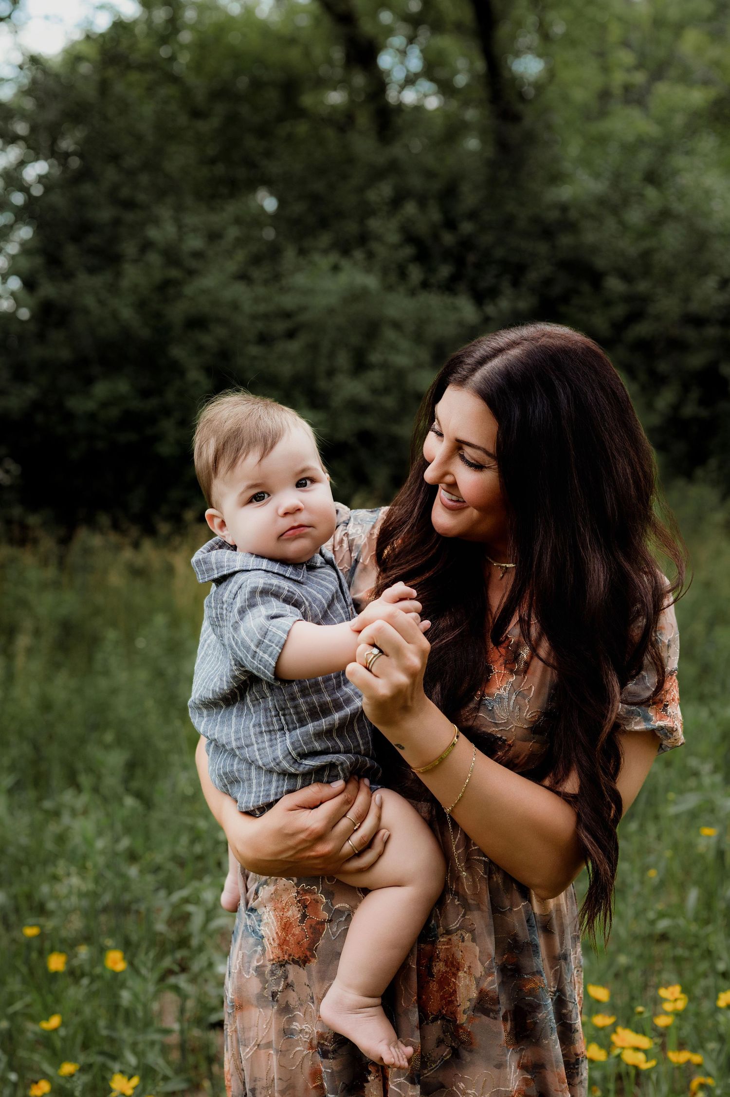A smiling mother with long dark hair holds her baby boy in a sunny wildflower meadow.