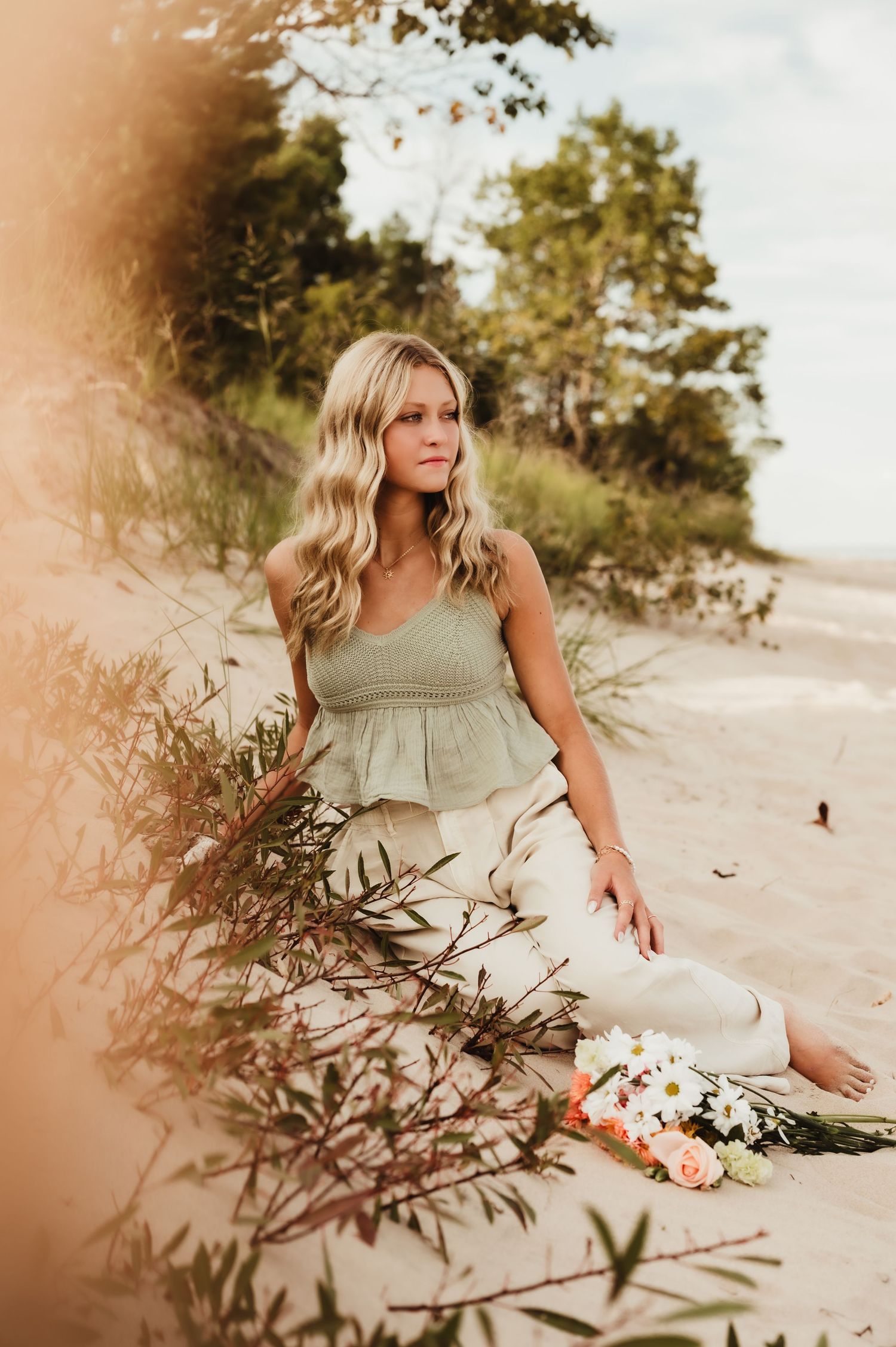 A woman in a sage green dress sits gracefully on a sandy beach with white flowers beside her in warm sunlight, Port Washington, WI