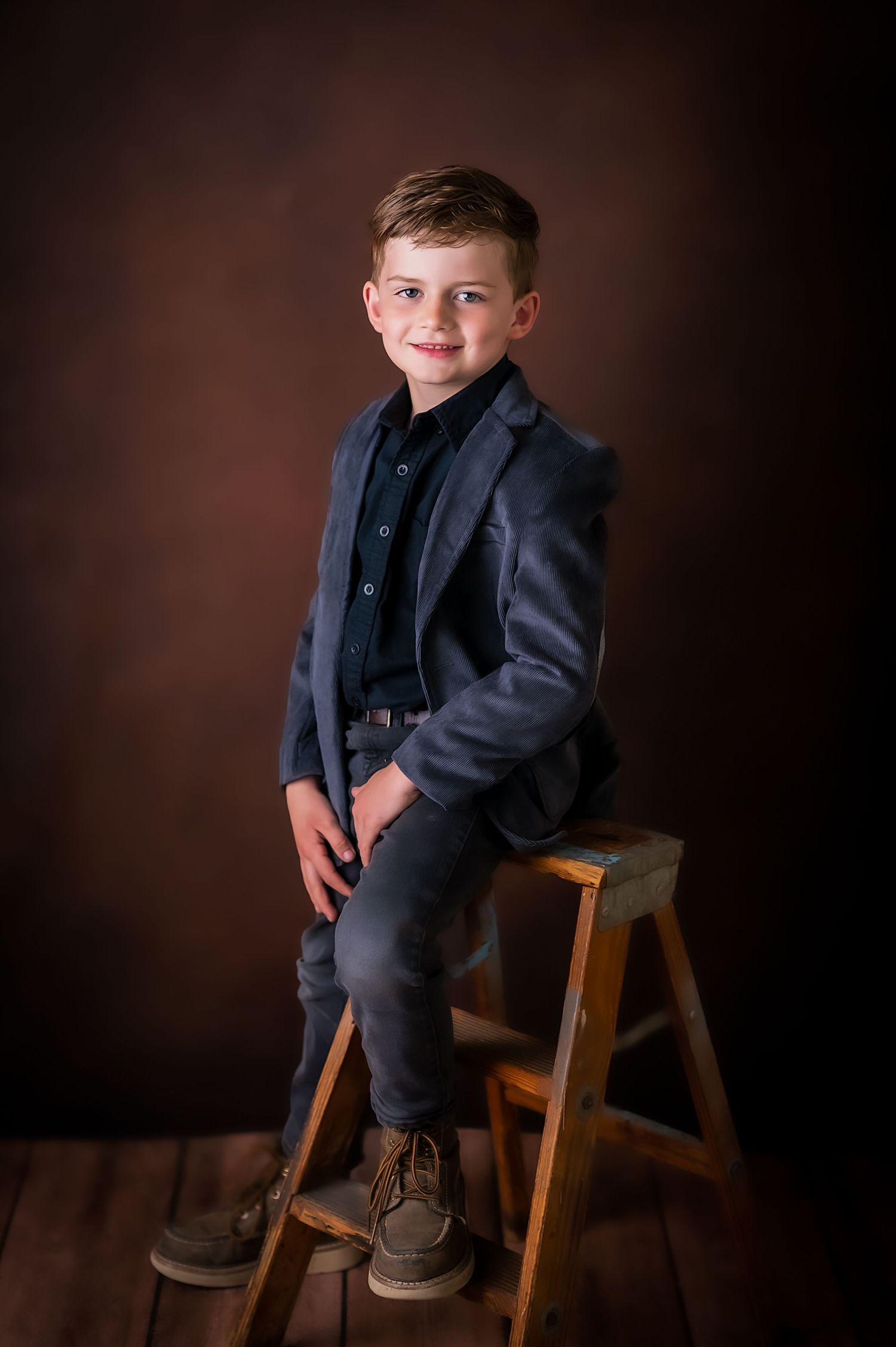 A young child in a dark suit sits casually on a wooden stool against a brown studio backdrop, Kewaskum, WI Studio