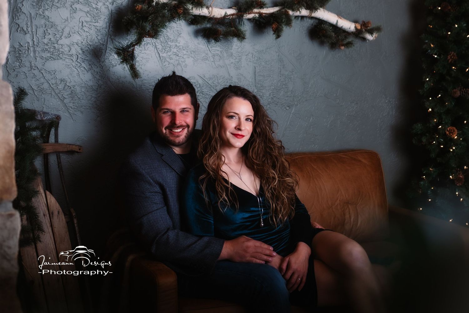Couple poses on brown leather couch in dimly lit room with holiday decorations.