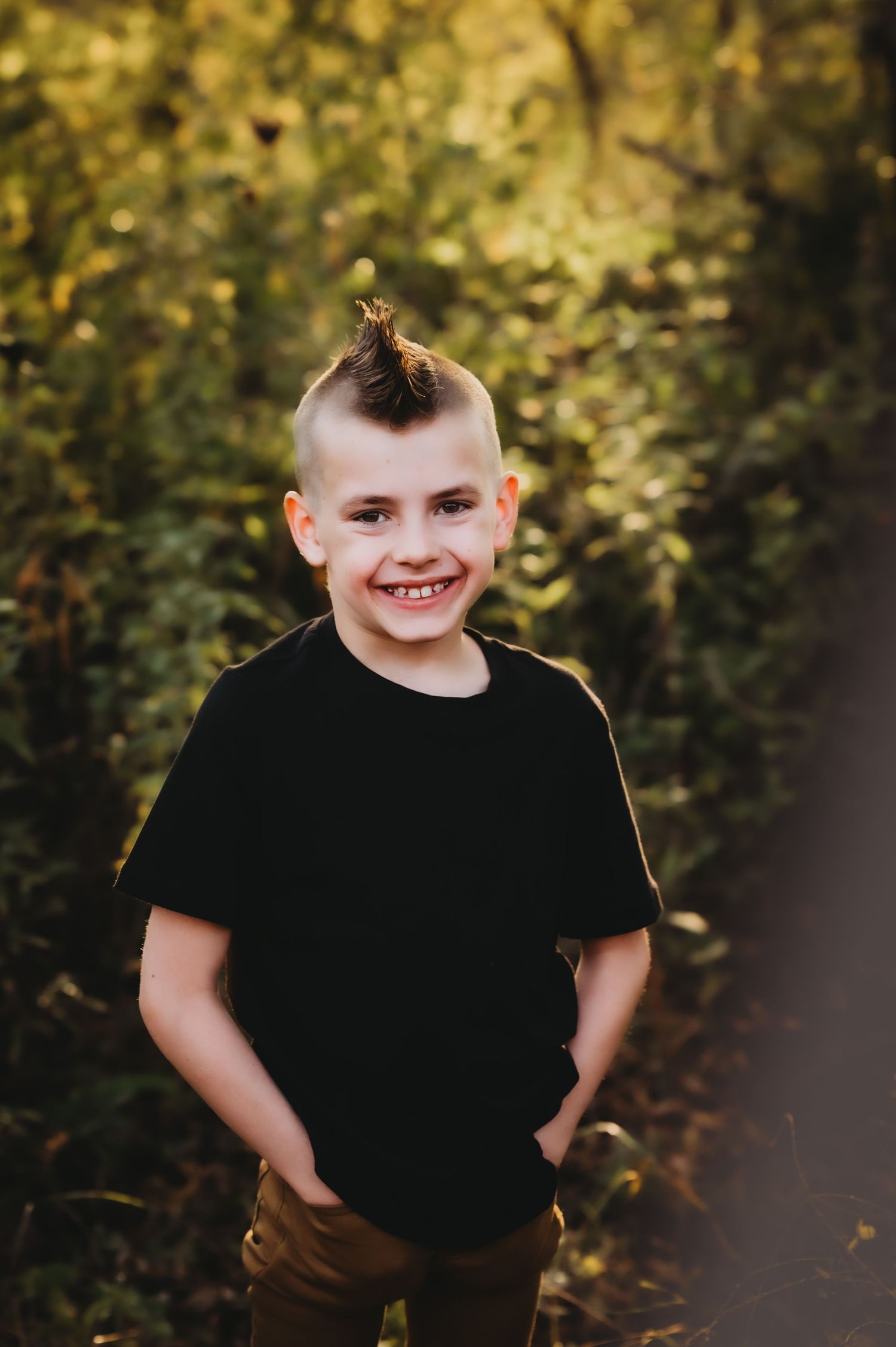 A child wearing a black t-shirt and brown pants stands outdoors against a blurred natural background.