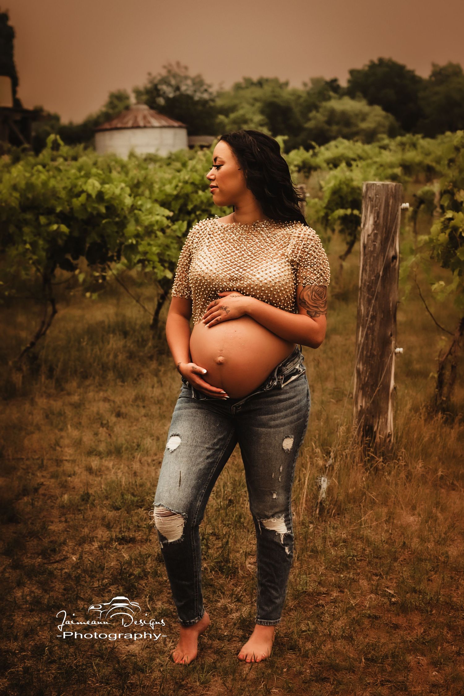 Barefoot maternity portrait in distressed jeans and sequined top against vineyard setting.