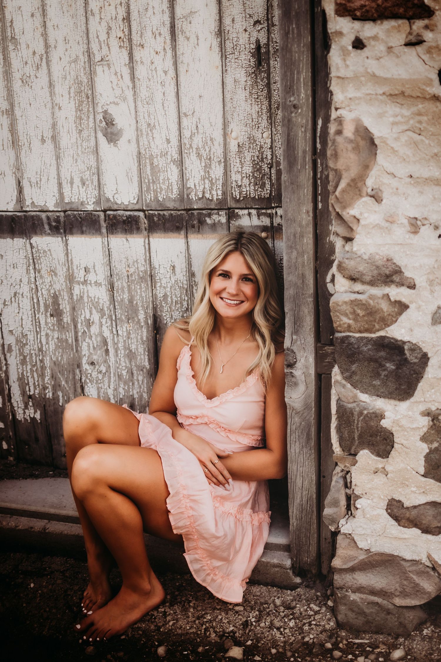 Person in pink dress sits by weathered wooden door and stone wall.