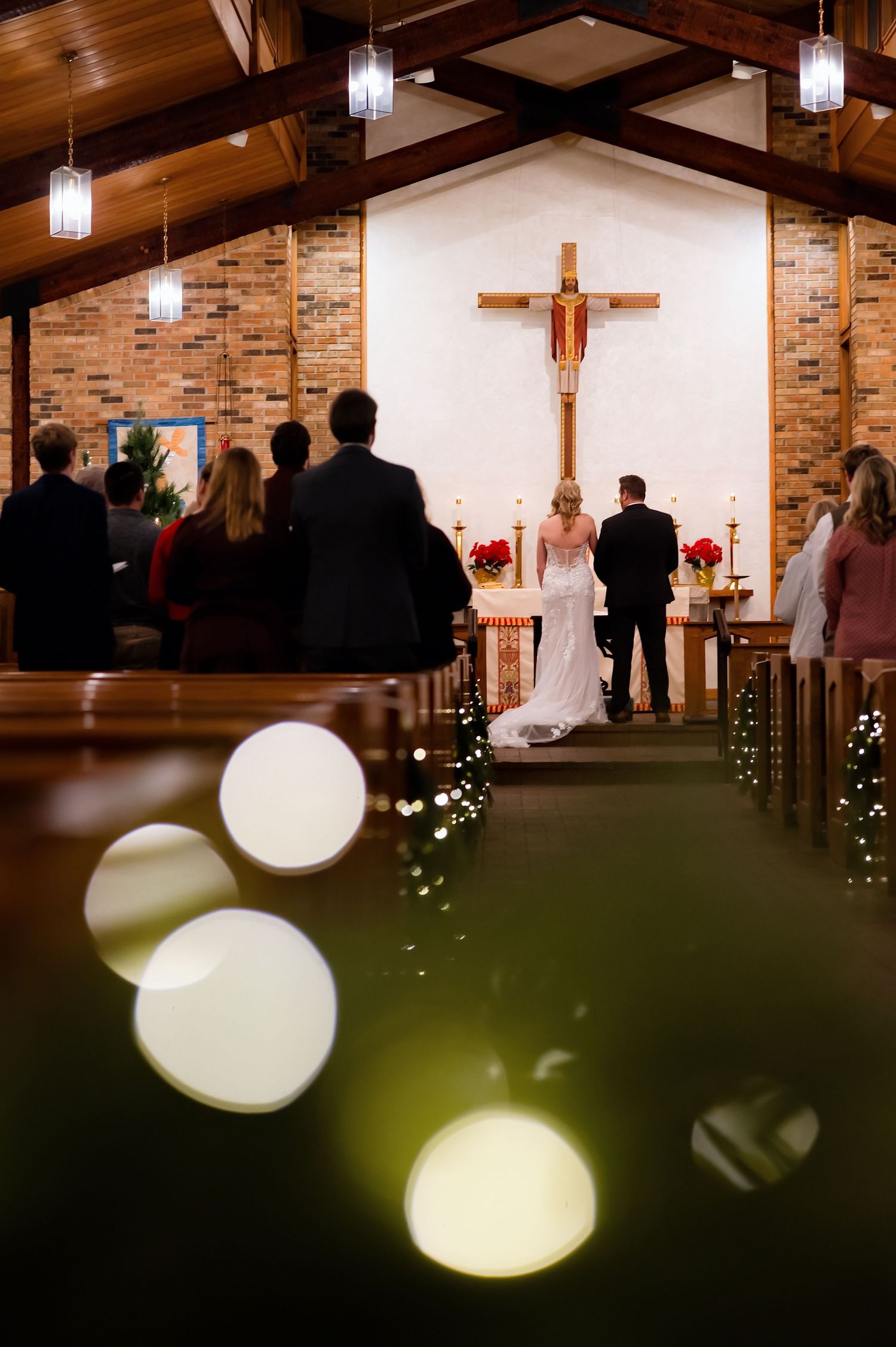 A wedding ceremony takes place in a church with exposed wooden beams, brick walls, and a crucifix mounted at the altar.