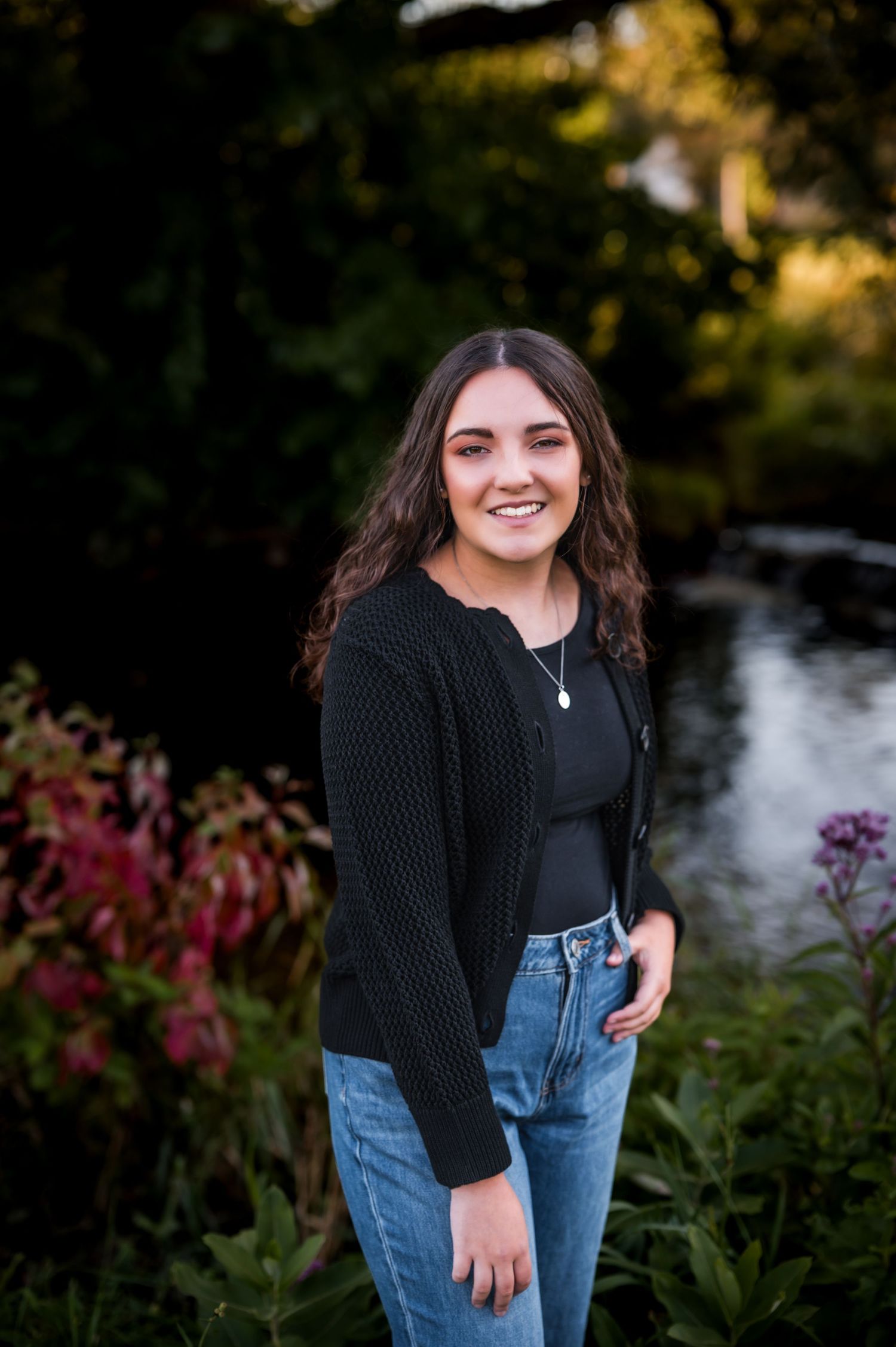 Portrait of someone in a black cardigan and jeans standing by a river at sunset with autumn foliage in the background., Kewaskum, WI