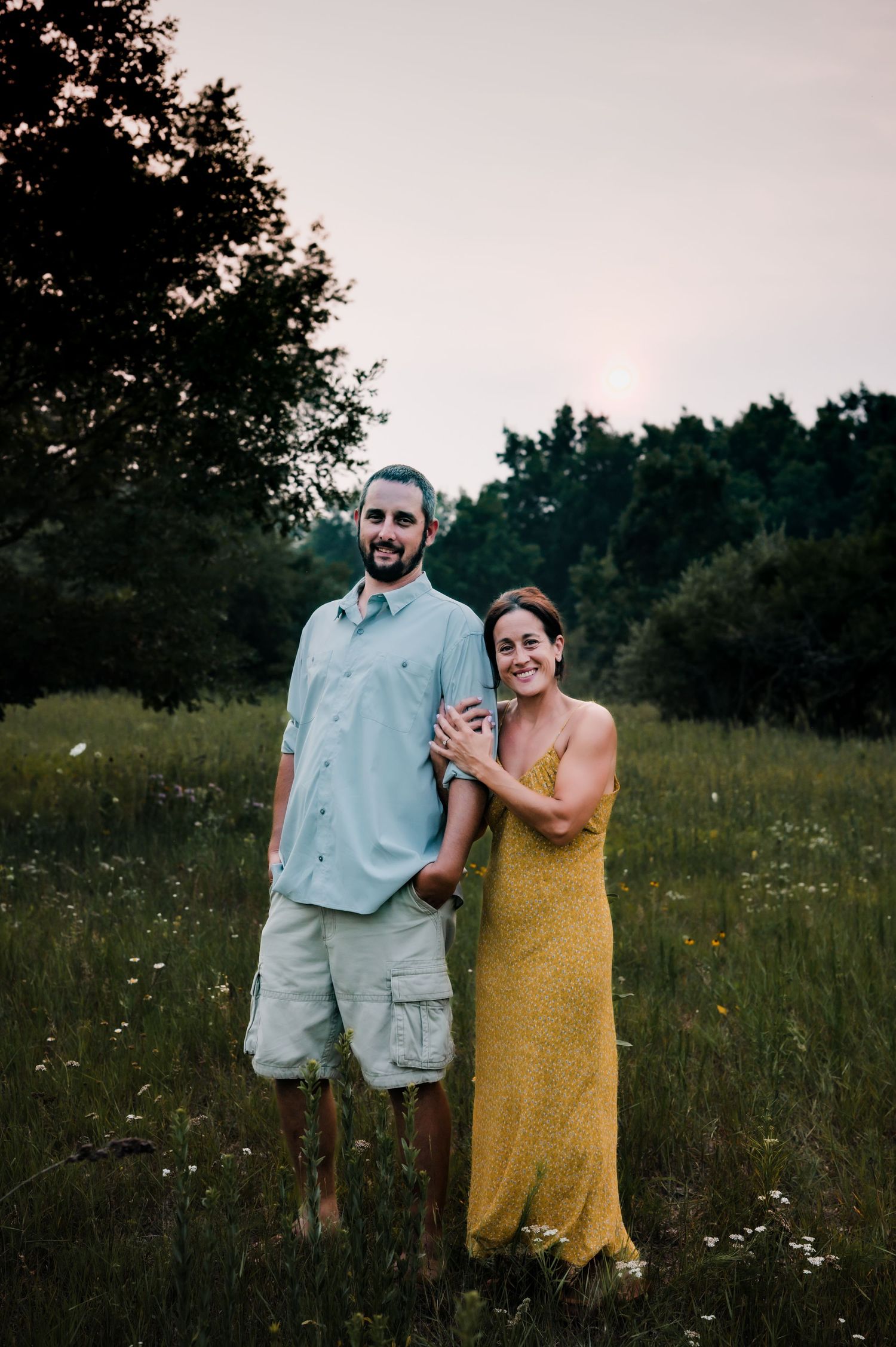 Couple smiling together in a sunny meadow, man in light blue shirt and shorts, woman in yellow dress, trees in background.