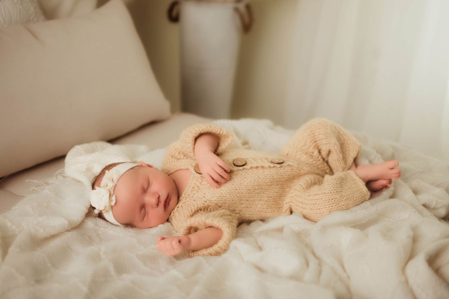 Newborn baby in a cream knit romper and white bow headband sleeping peacefully on a soft white blanket with a neutral pillow nearby.