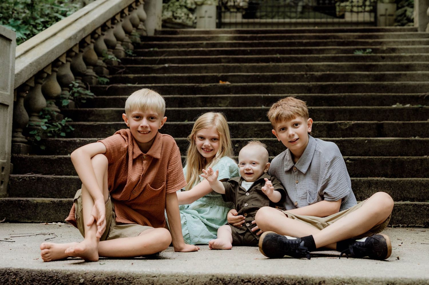 Four young siblings pose together on stone steps outdoors, smiling for a family portrait photo.