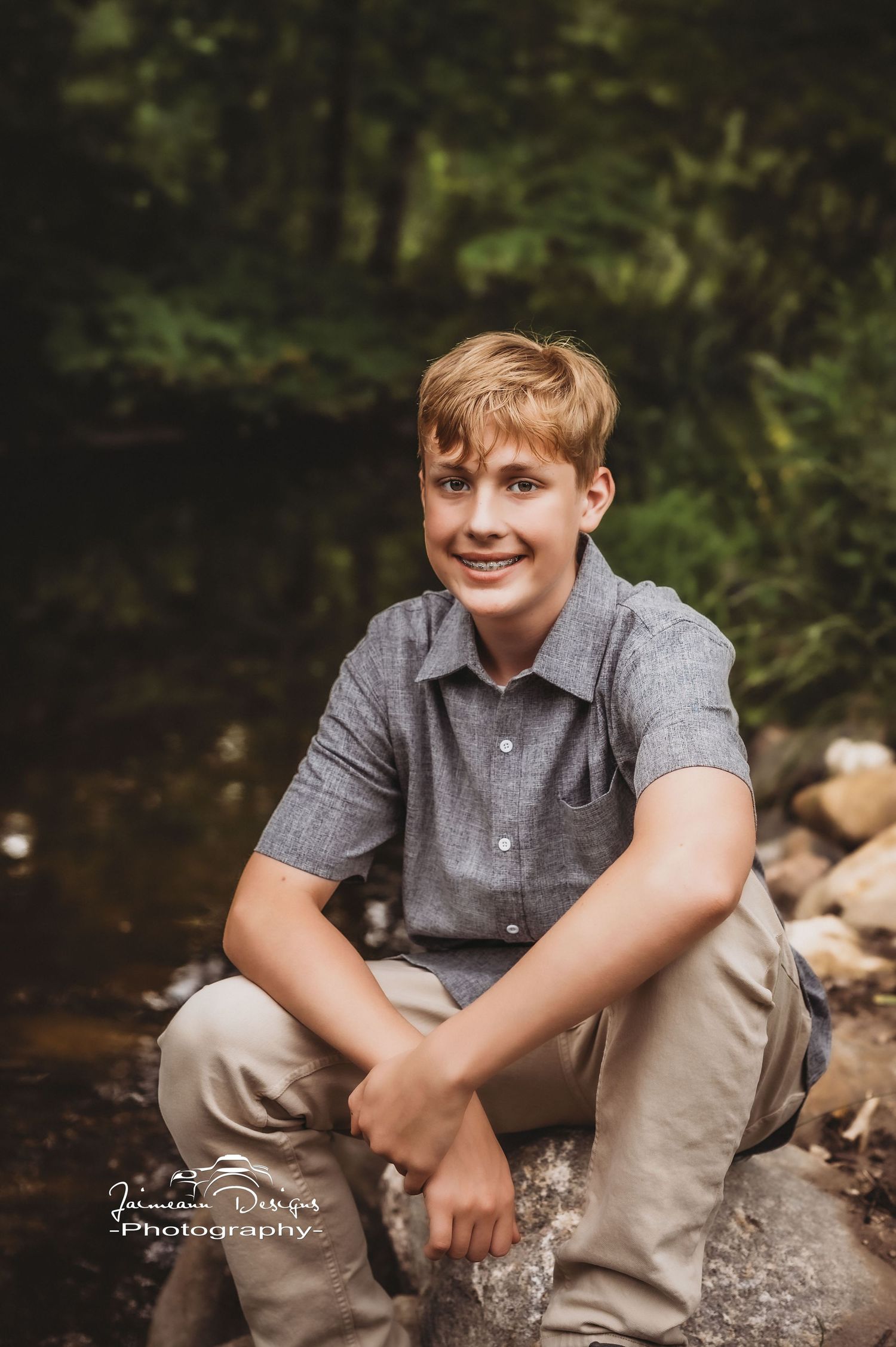 An outdoor portrait of someone sitting on rocks by a stream wearing casual grey attire.