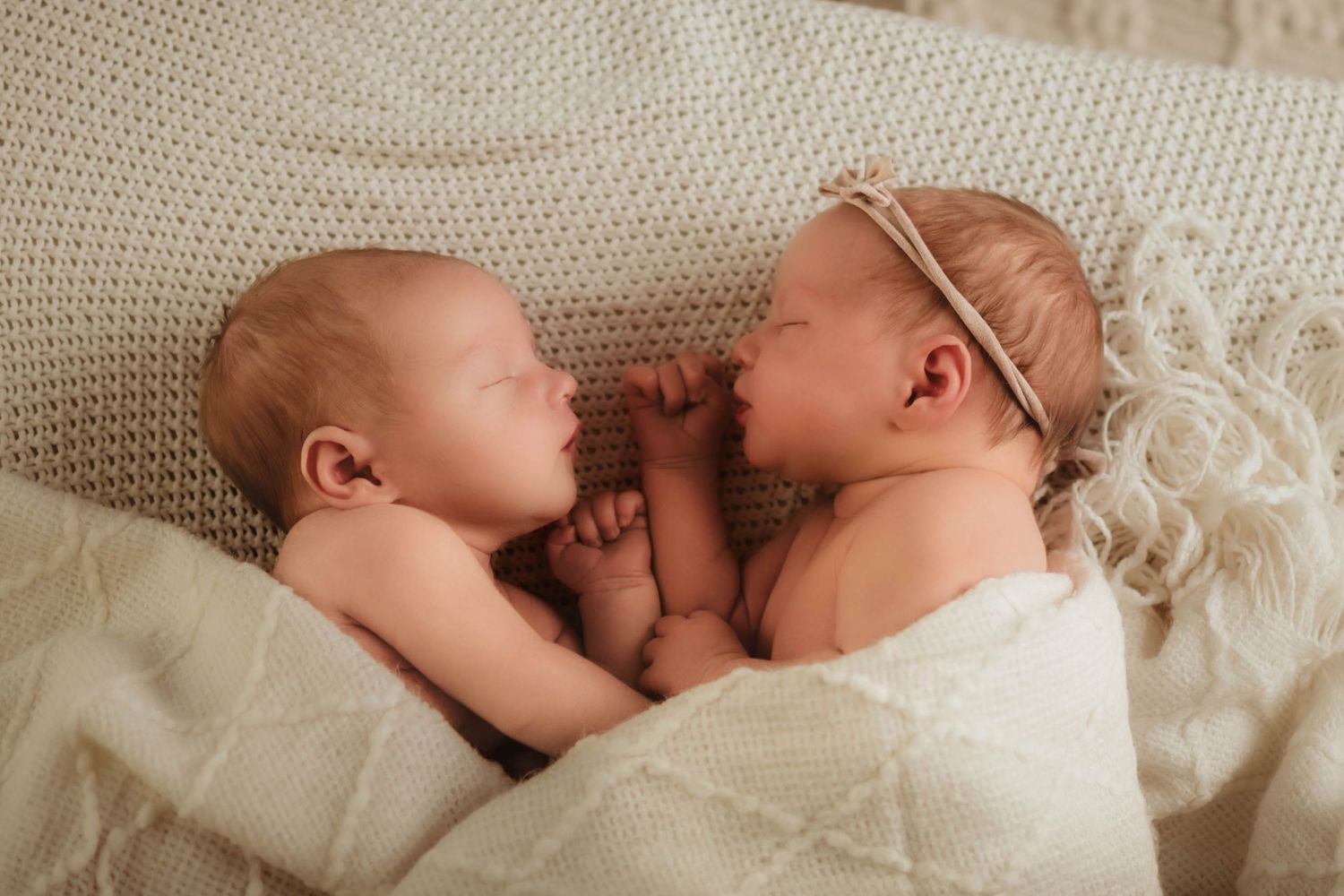 Two sleeping newborn babies lying side by side on a cream knit blanket, one wearing a delicate headband, with hands touching.