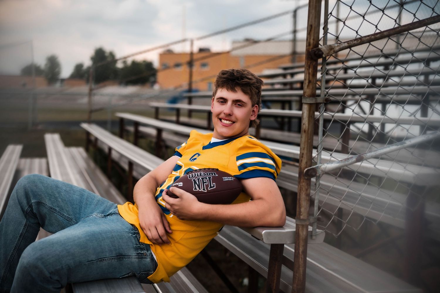 Young male football player in yellow jersey holding ball, sitting relaxed on bleachers at outdoor stadium.