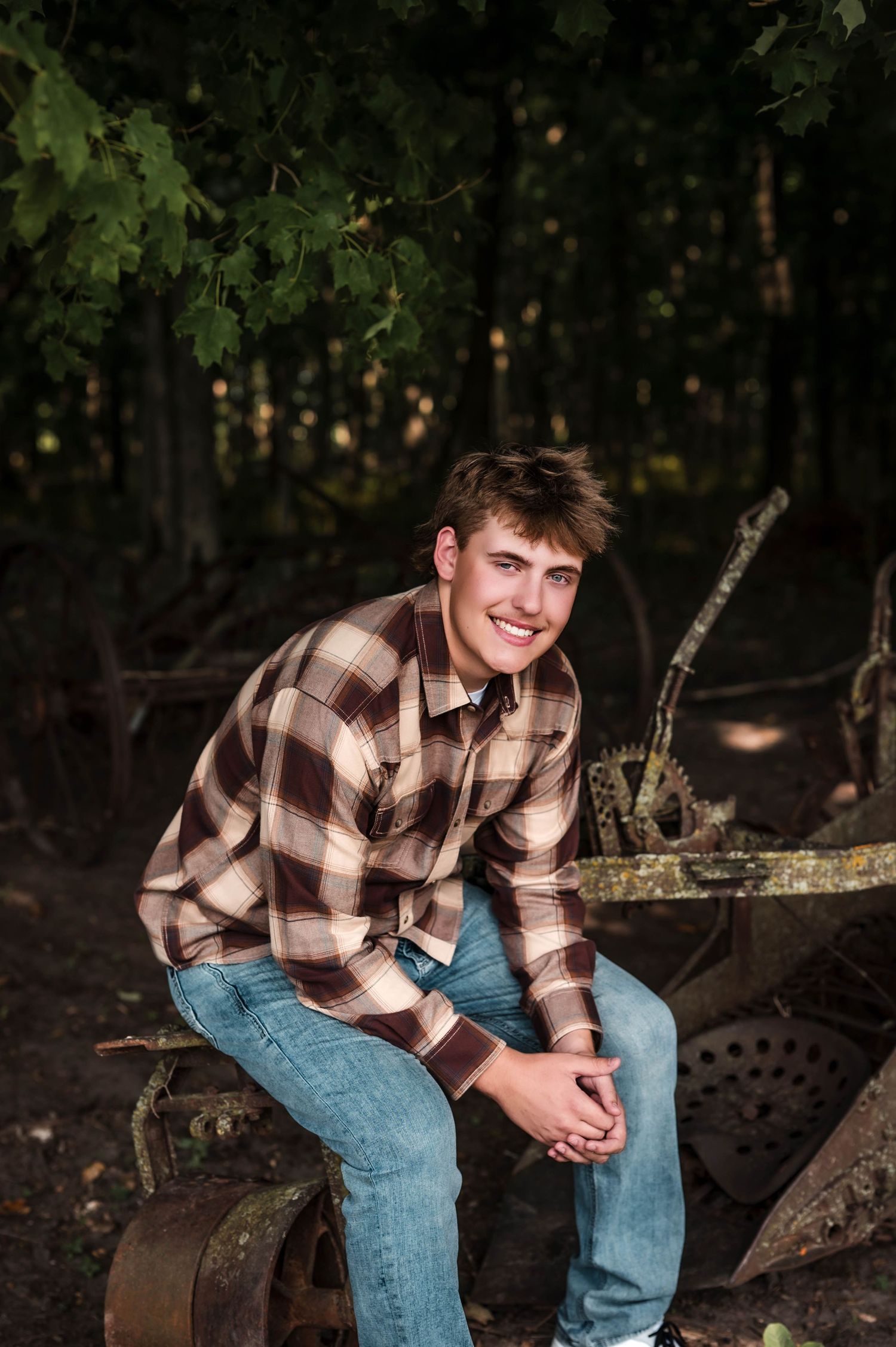 Young man in plaid flannel shirt and jeans sits smiling on rustic farm equipment surrounded by lush green trees.