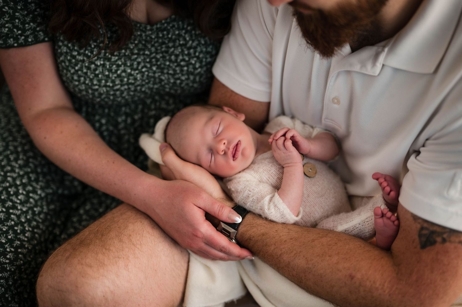 Parents gently cradle a sleeping newborn baby wrapped in a soft cream outfit during an intimate, tender family photo session.