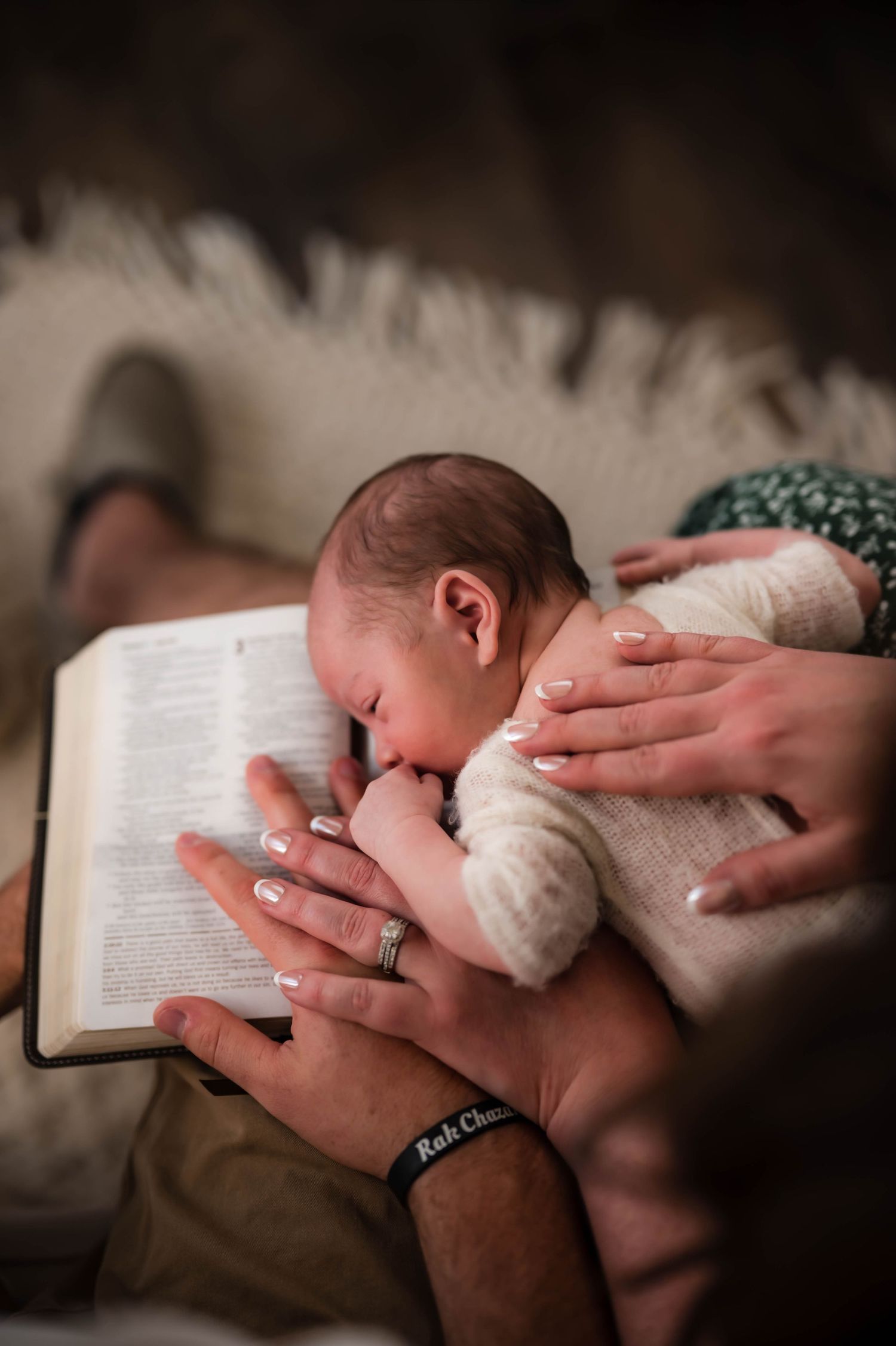 Newborn baby resting its head on an open Bible while parents' hands gently cradle the sleeping infant in a tender family moment.