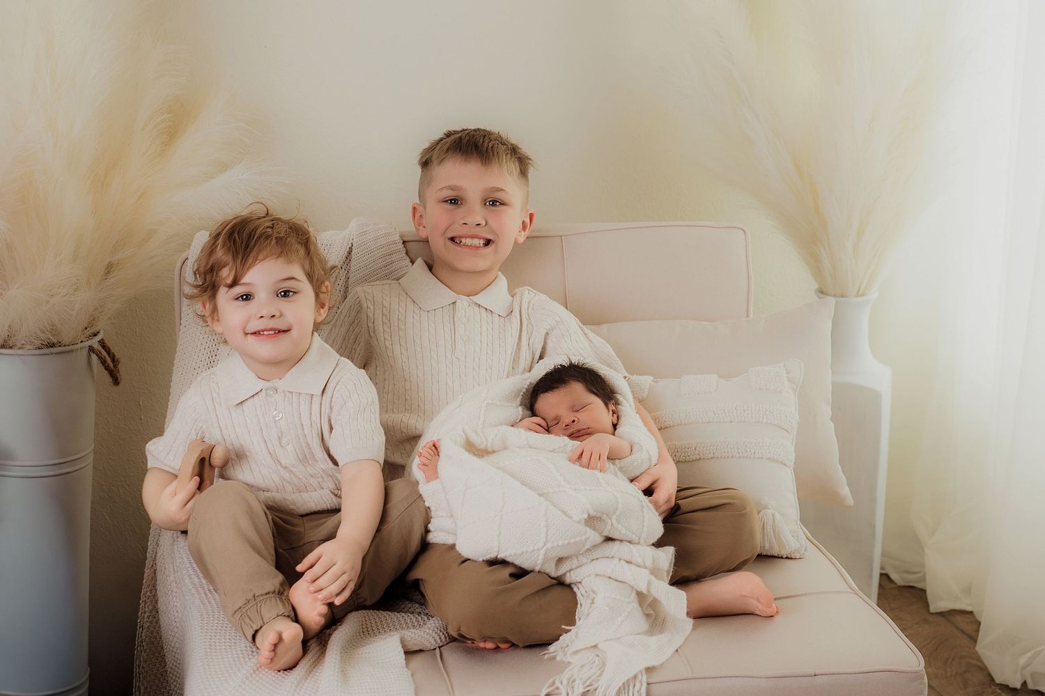 Two young boys and a newborn baby sibling pose together on a beige chair in a soft, neutral-toned studio setting.