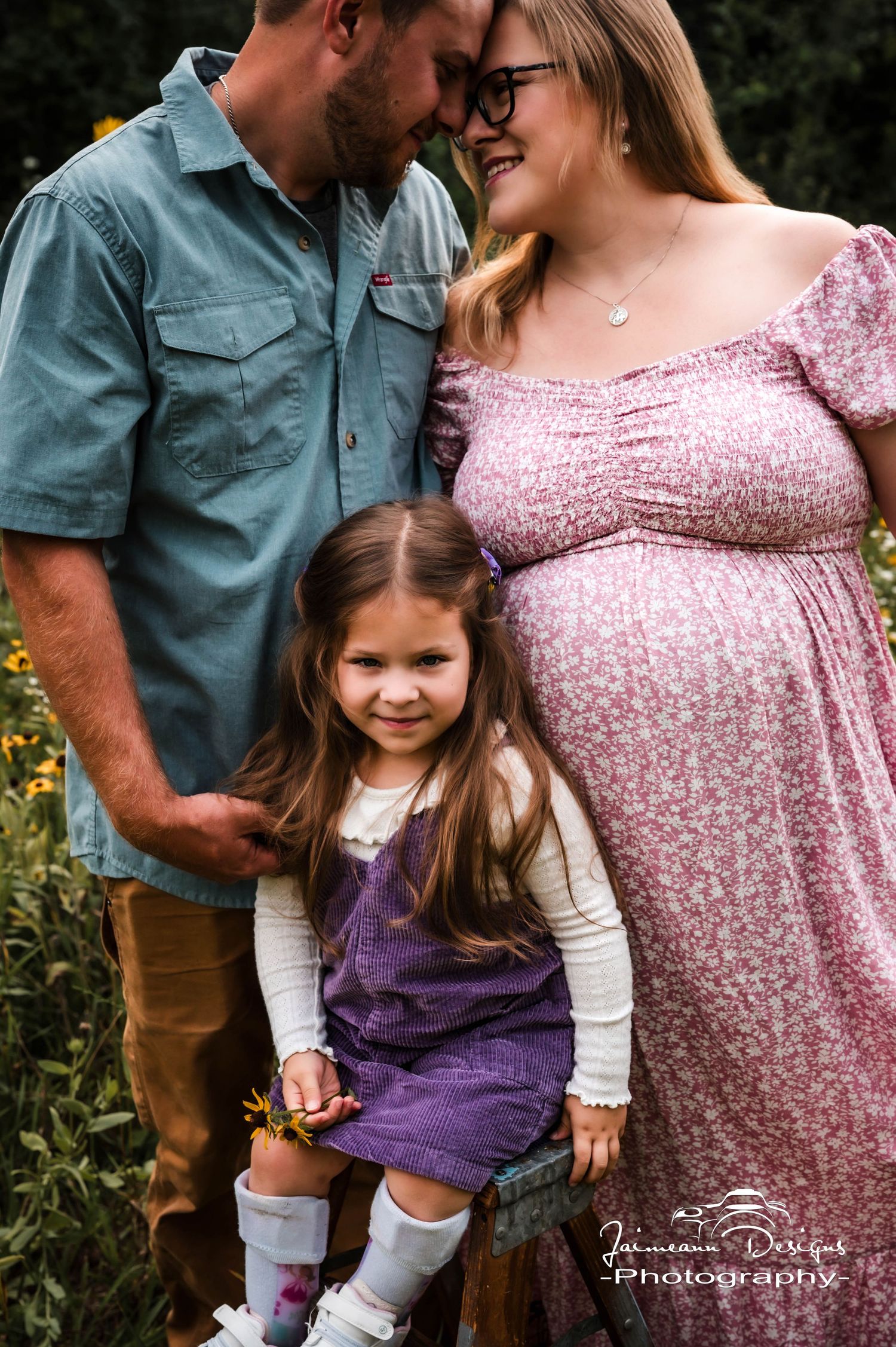 Pregnant woman in pink floral dress with husband and young daughter in purple outfit posing outdoors among wildflowers.