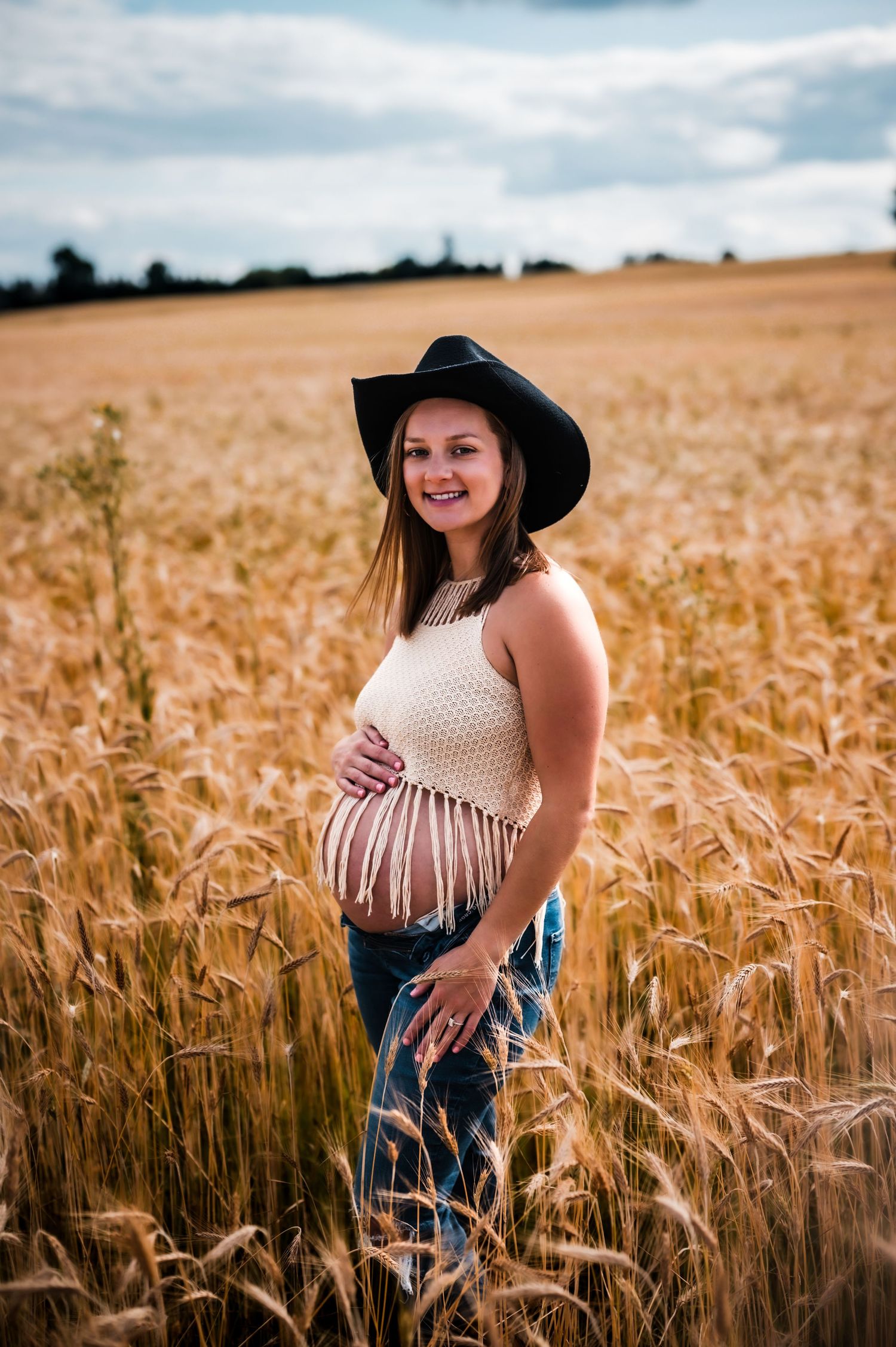 Pregnant woman in cowboy hat and crochet top holding belly in golden wheat field.