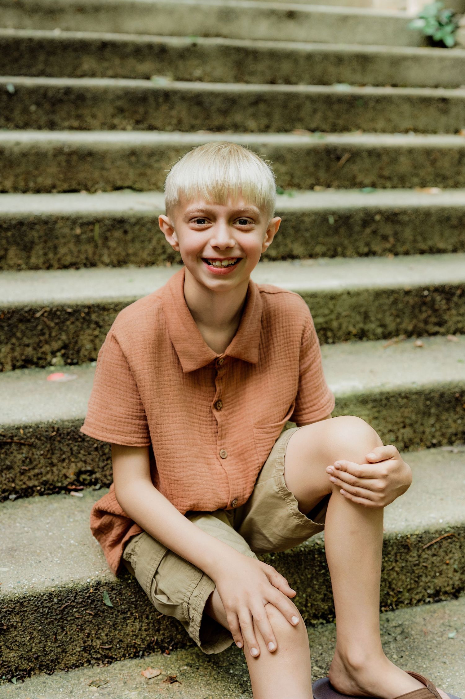 Smiling blonde boy in rust shirt and khaki shorts sitting on stone steps outdoors.