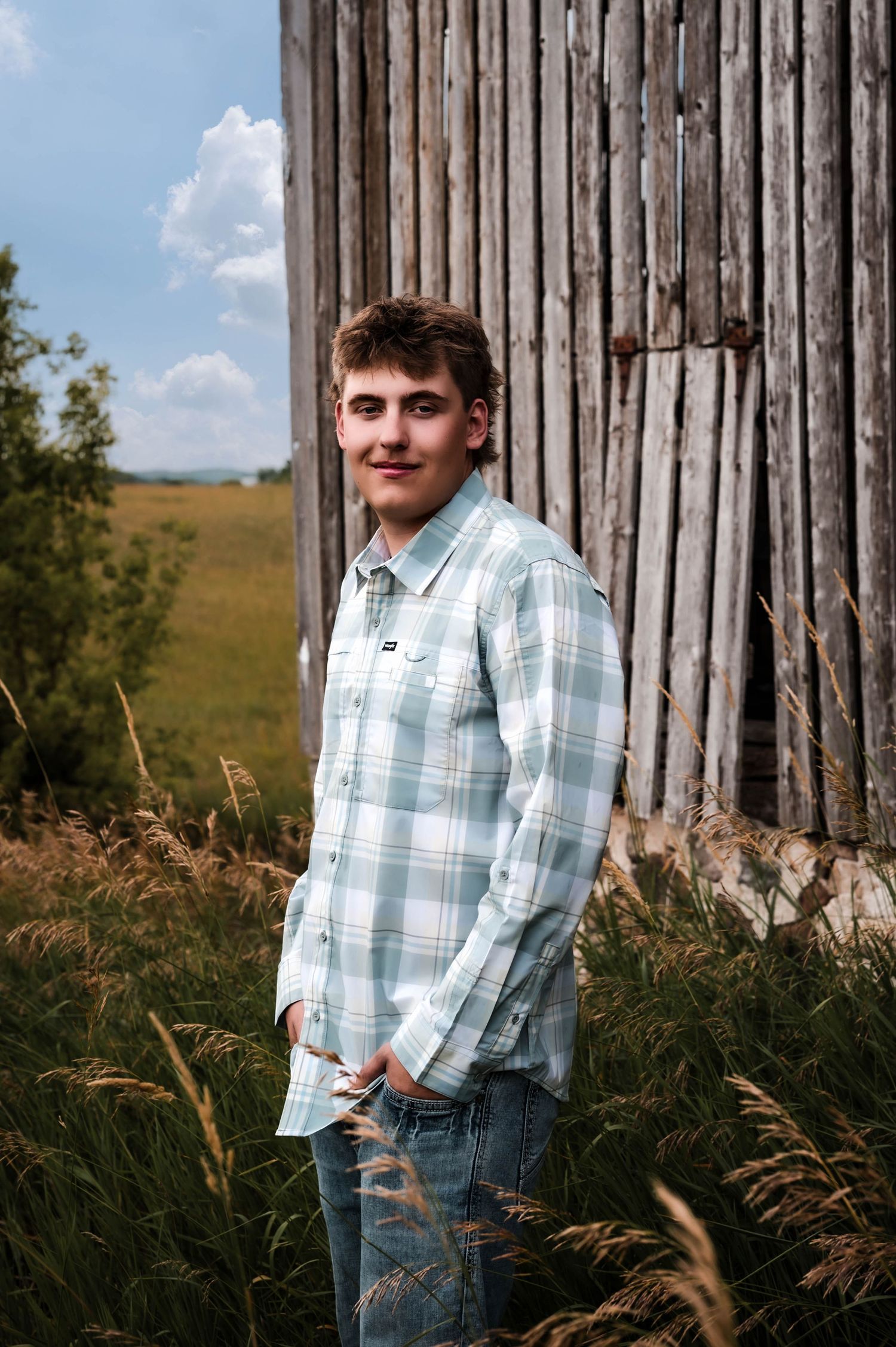 Young man in plaid shirt and jeans smiling beside a rustic wooden barn in a green rural field.