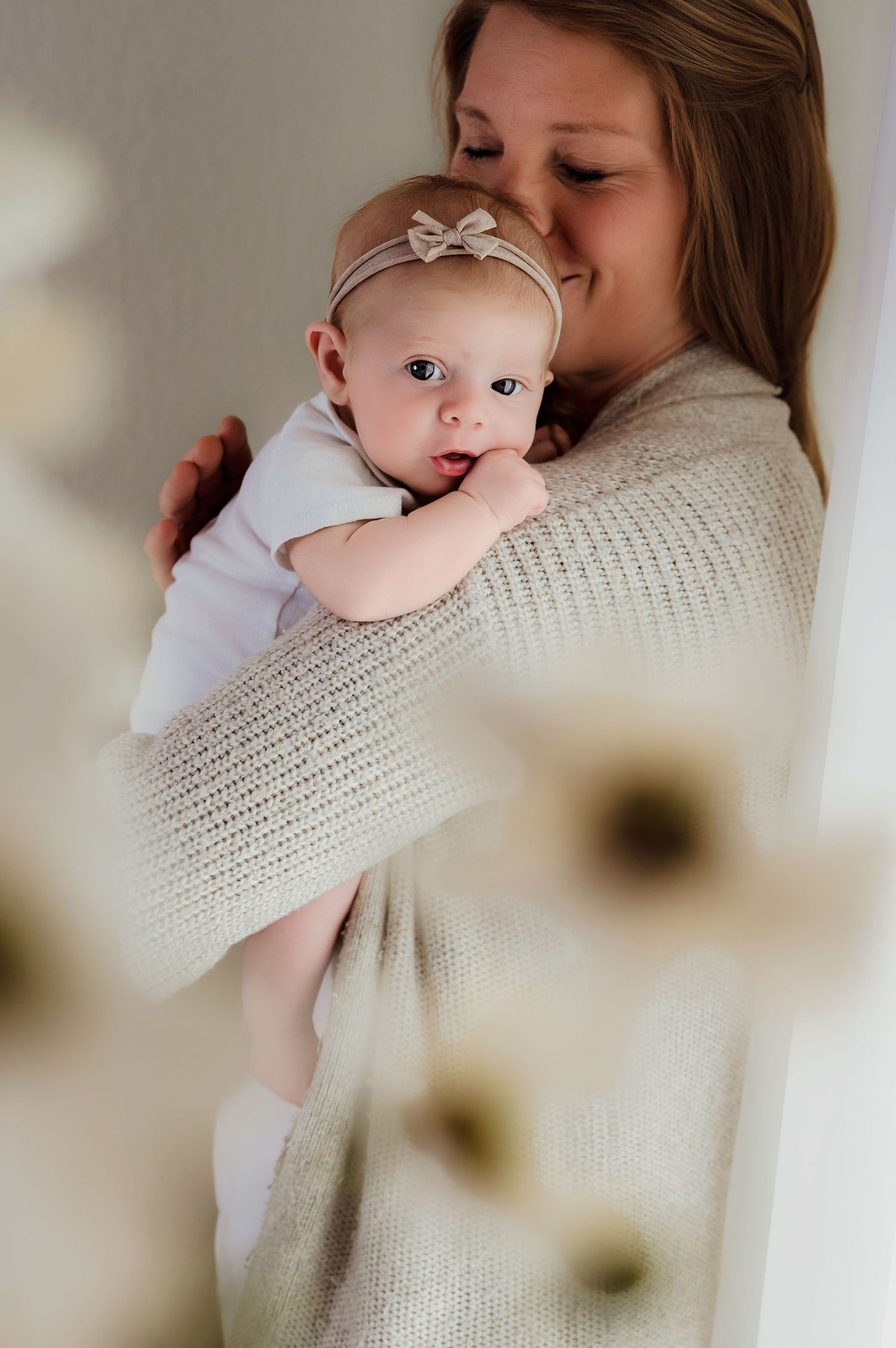 A smiling mother in a cream knit sweater tenderly holds her blue-eyed baby girl wearing a bow headband.