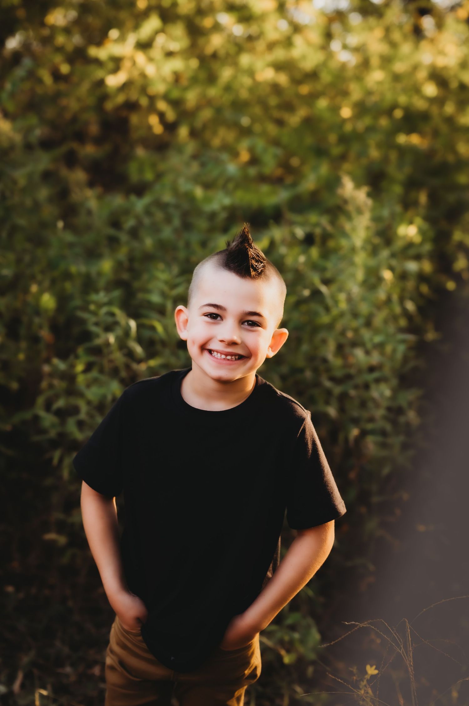 Child wearing black t-shirt and brown pants stands smiling outdoors with autumn foliage in background.